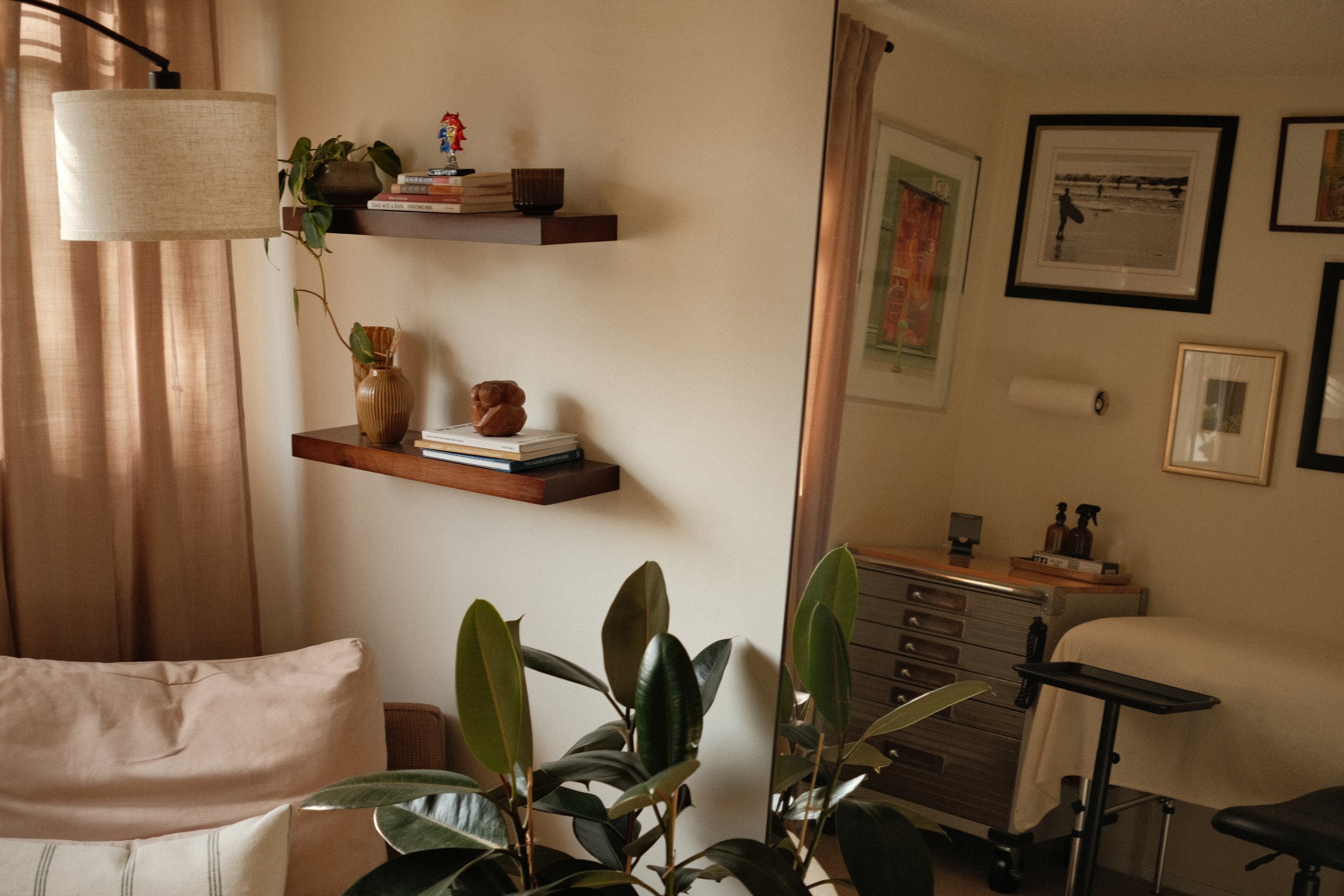 A cozy living room corner with a beige sofa, green houseplants, shelf with books and small ornaments, framed pictures on the wall, a dresser, and a mirrored surface reflecting the room.