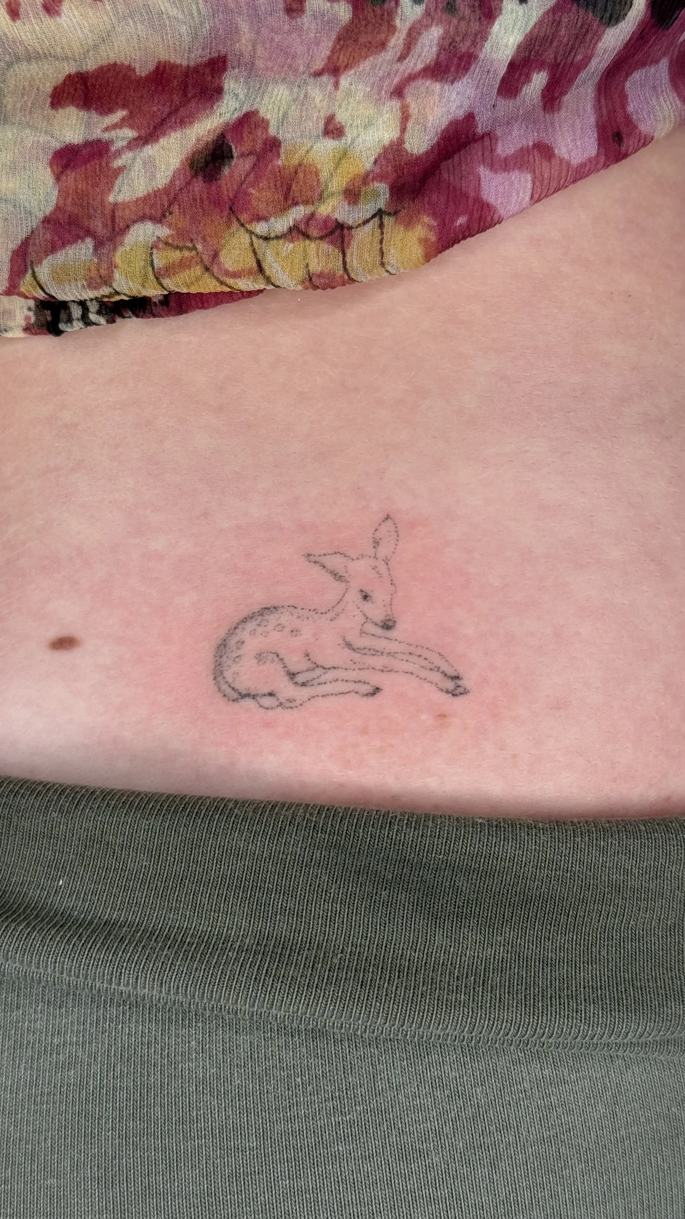 A close-up of a person's skin with a small elastic tattoo of a sitting deer with antlers, on the lower abdomen. The person is wearing a floral-patterned garment.