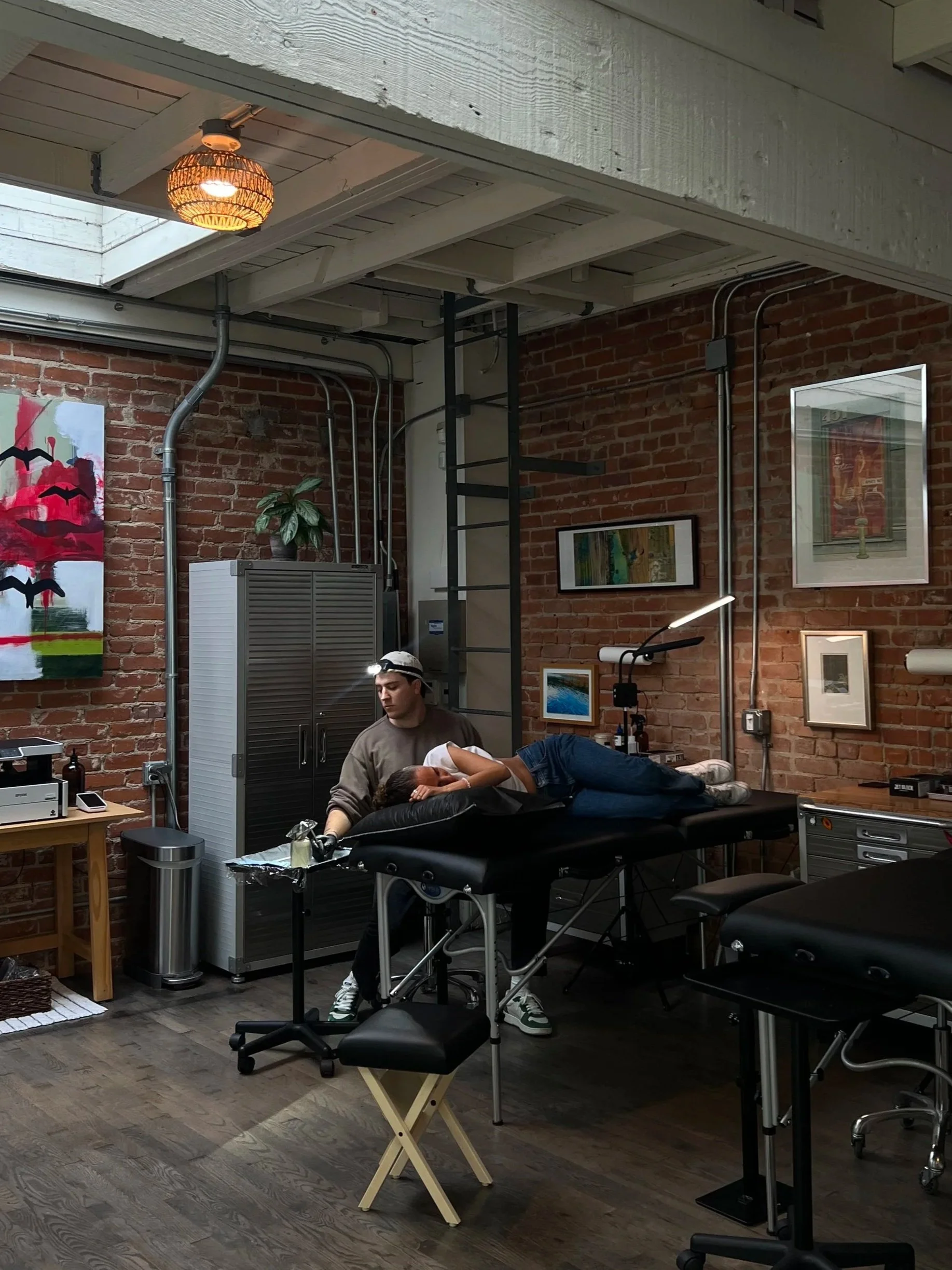 A person is lying on an examination table with a medical professional attending to them, in a room with exposed brick walls, framed artwork, and medical equipment.