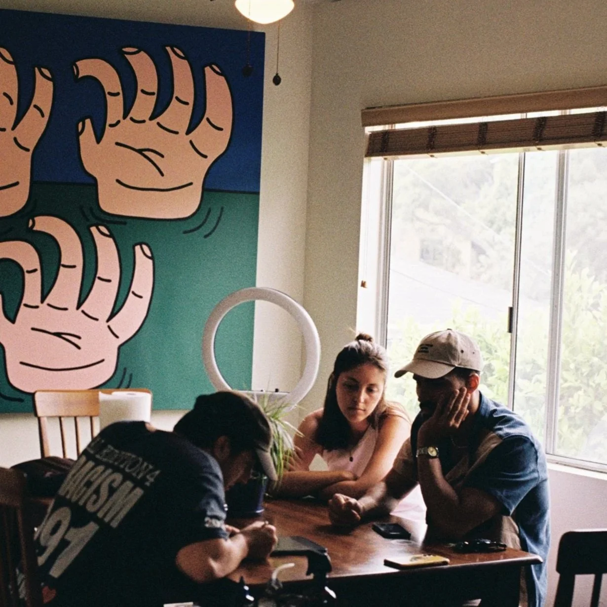 Three young adults sitting at a wooden table in a room with a large window and a colorful wall art of cartoonish hands making a thumbs-up gesture. One person is wearing a cap, and two are focused on their phones.