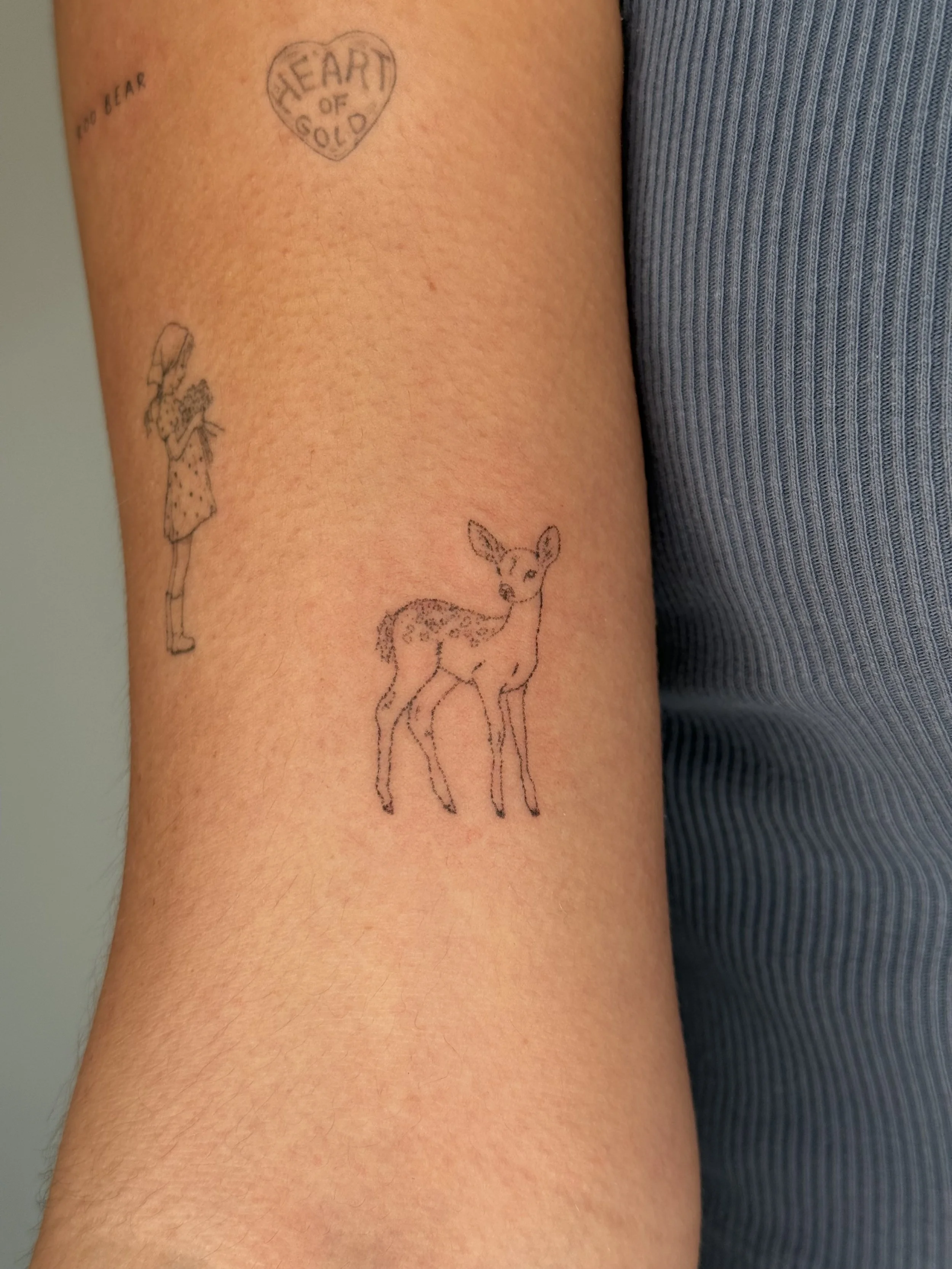 A person's upper arm with four tattoos, including a heart-shaped tattoo with the words "HEART OF GOLD," a small tattoo of a girl holding a flower bouquet, a small tattoo of a deer, and the words "NO FEAR" in script.