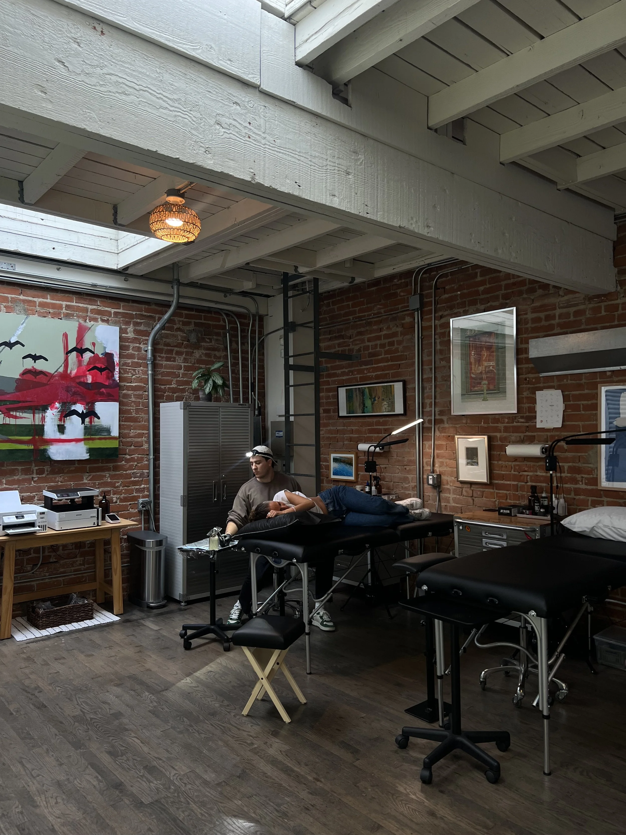A person lying on a medical examination table with a healthcareworker attending to them in a room with brick walls, artwork on the walls, and medical equipment.