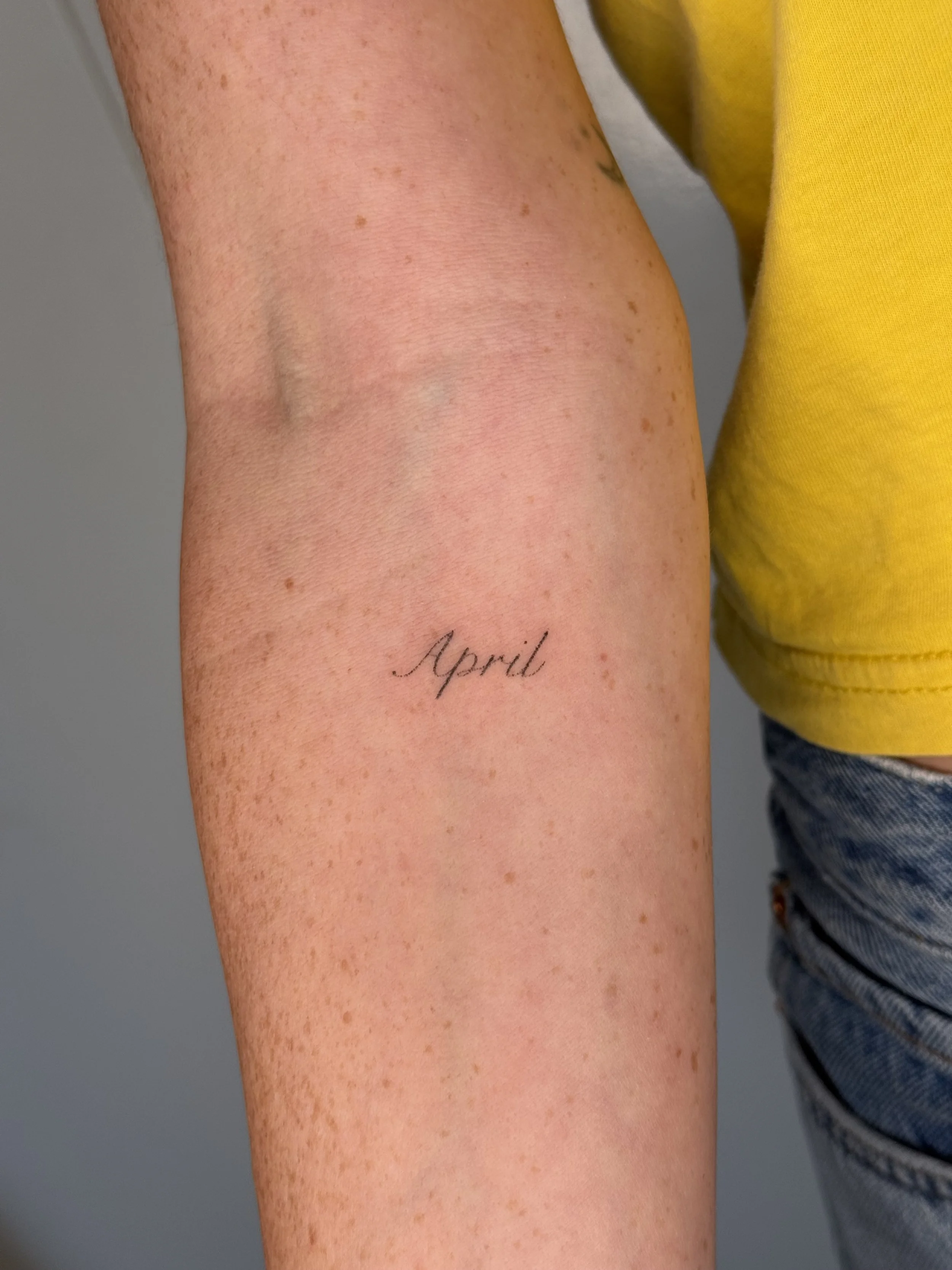 Close-up of a person's arm with freckles and a tattoo that reads 'April' in cursive.