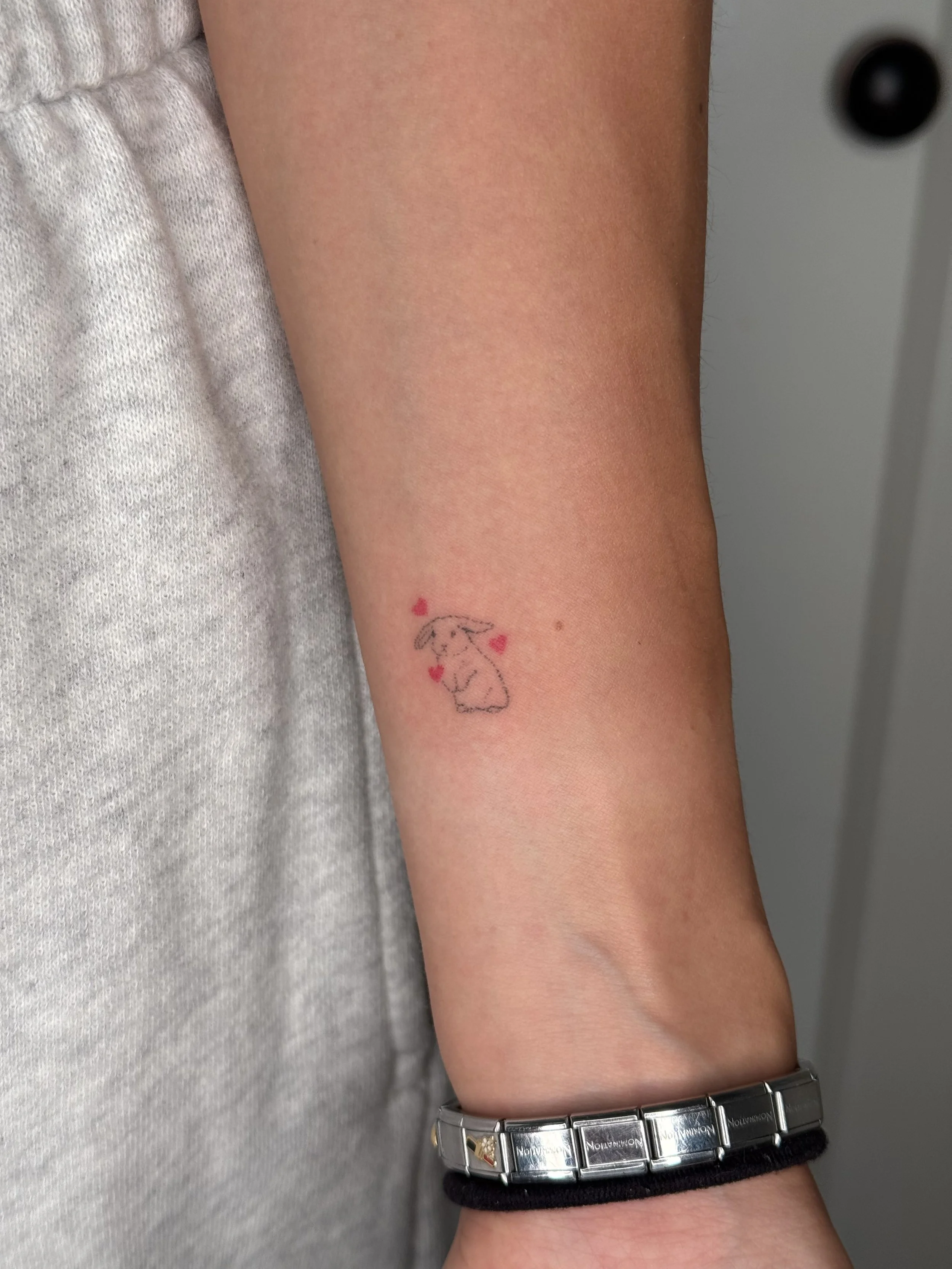A person's arm with a small bunny and heart tattoo, wearing a metallic and black bracelet, next to a gray sleeve.