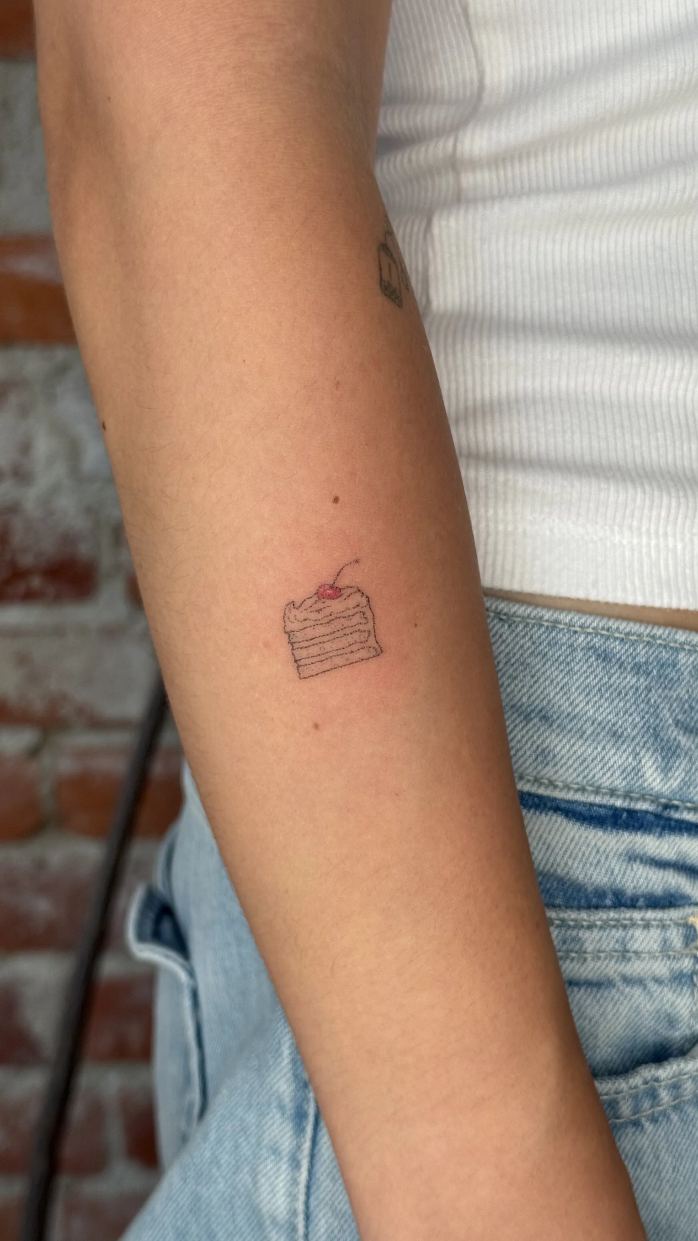 Small tattoo of a slice of cake with a cherry on top on the person's forearm.
