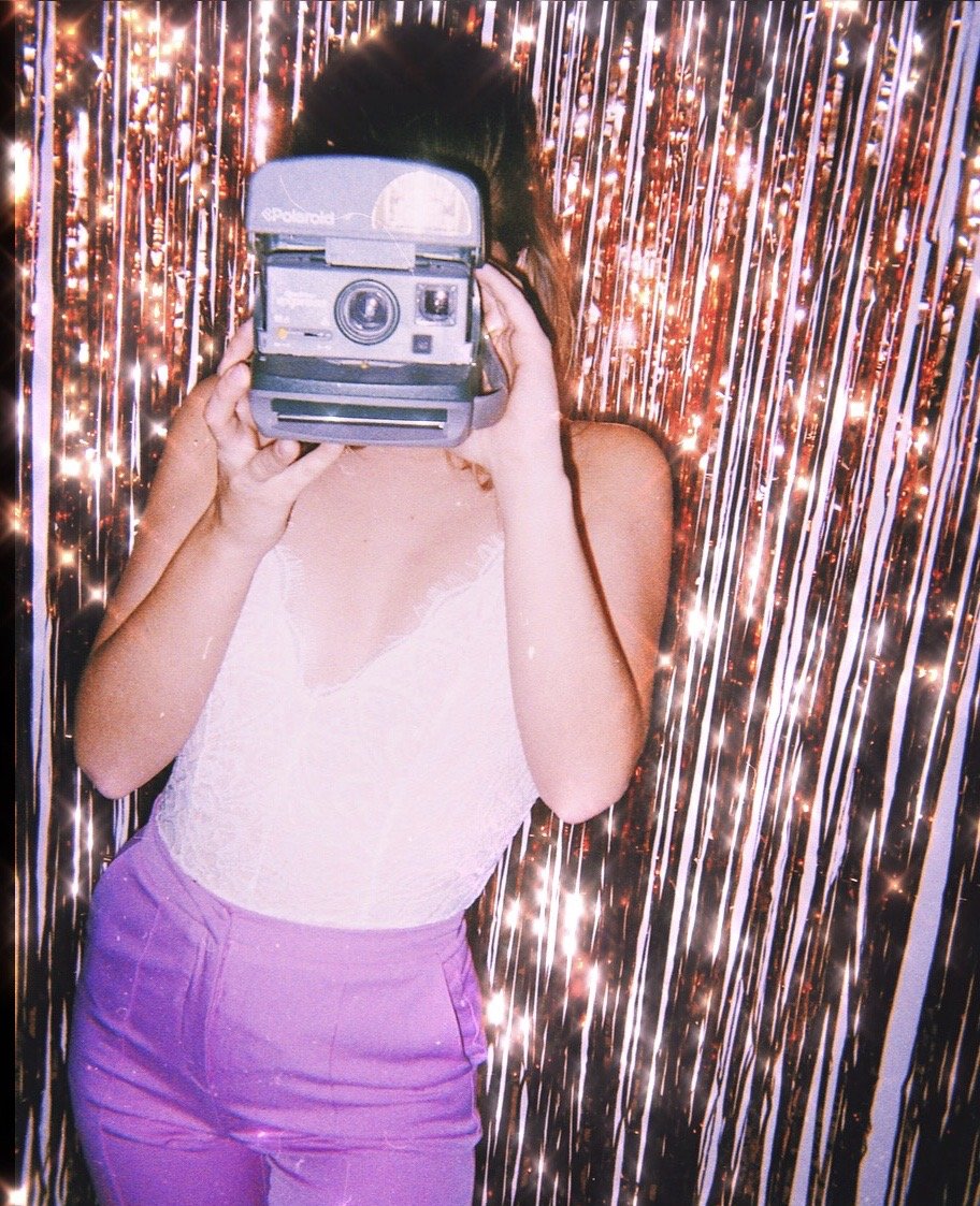 A person taking a photo with a Polaroid camera in front of a shimmering, sparkly backdrop.