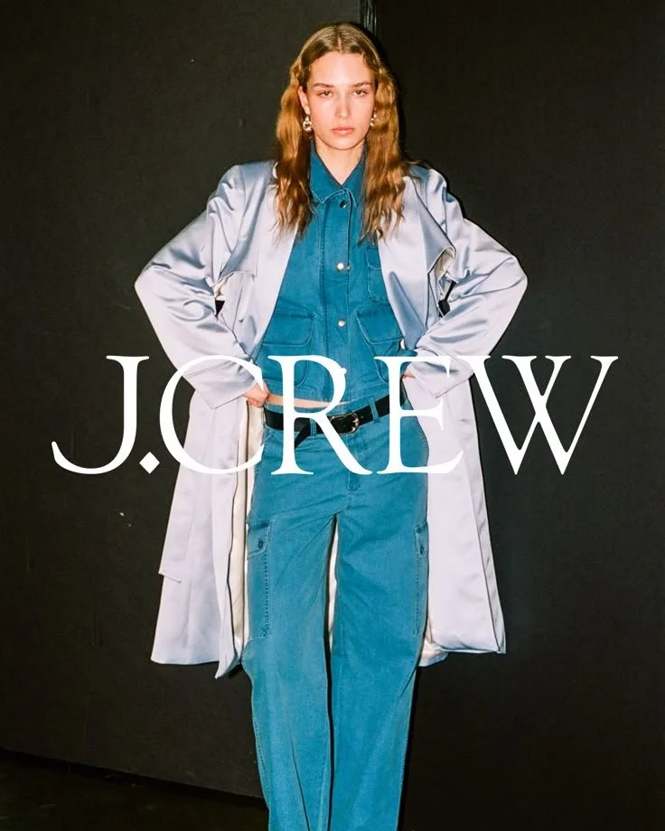 Young woman with long wavy hair wearing a blue denim shirt and jeans, a white jacket, standing with hands on hips in front of a black background, with 'J.CREW' text overlay.