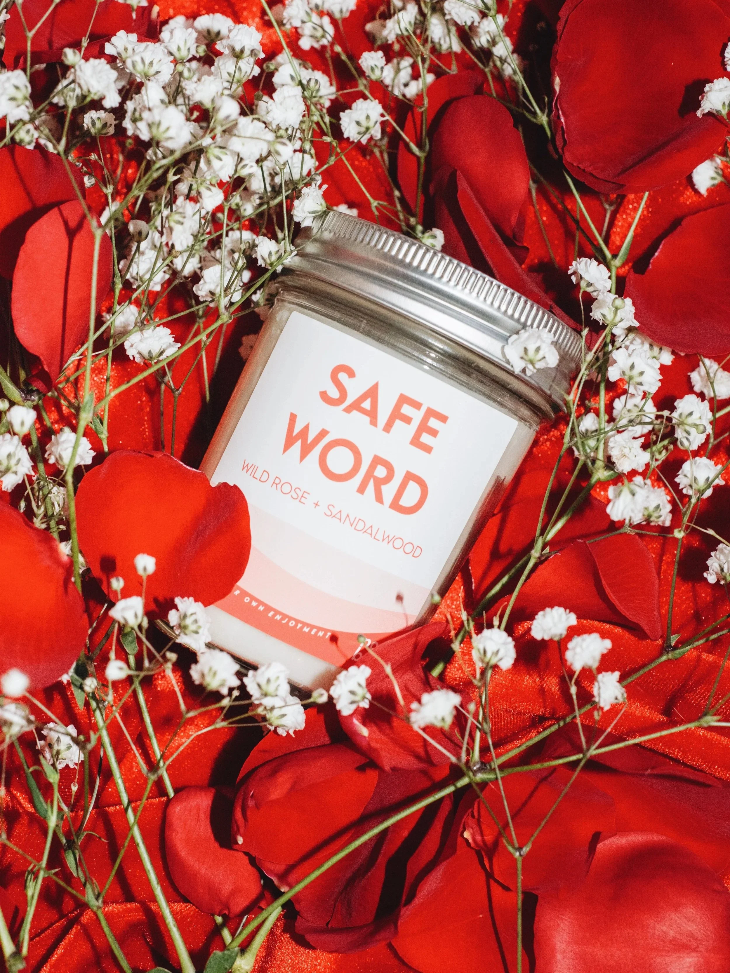 A candle labeled 'SAFE WORD' surrounded by red rose petals and small white flowers.
