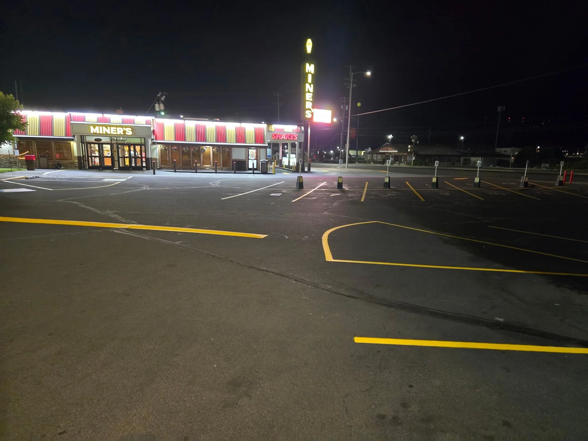Empty fast food restaurant parking lot at night with illuminated signs for Miner's and Shakes, and neon sign for Miner’s.