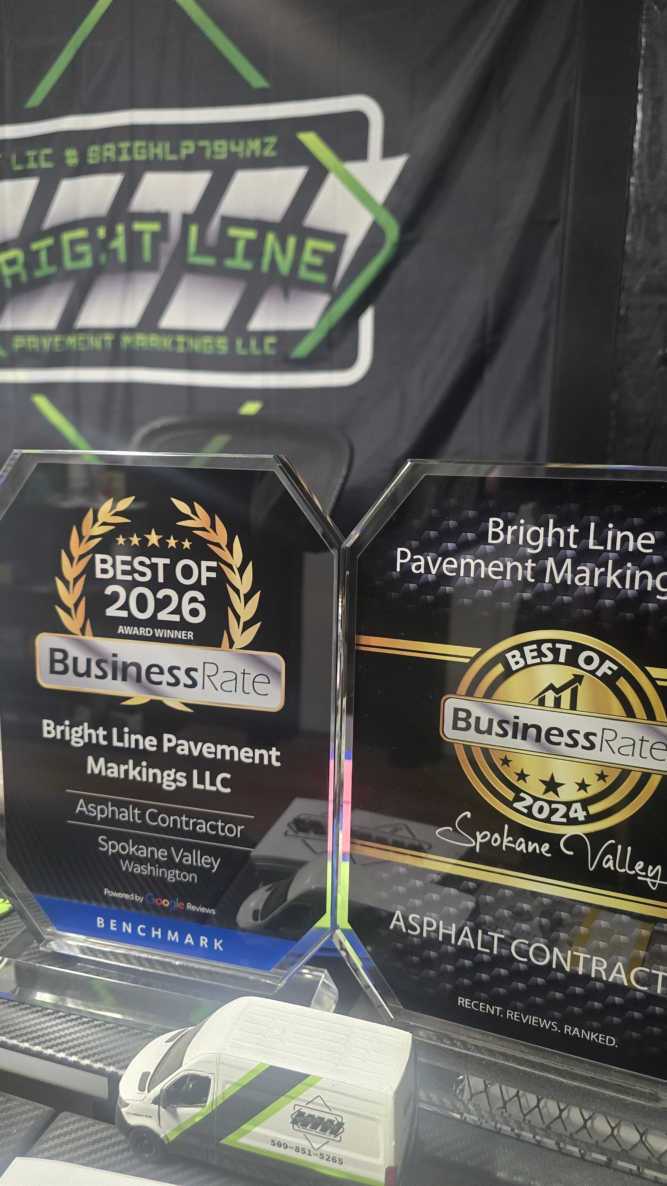 Two award plaques for Bright Line Pavement Markings LLC, recognizing as Best of 2026 and 2024 by BusinessRate, with signs indicating asphalt contractor services in Spokane Valley, Washington.
