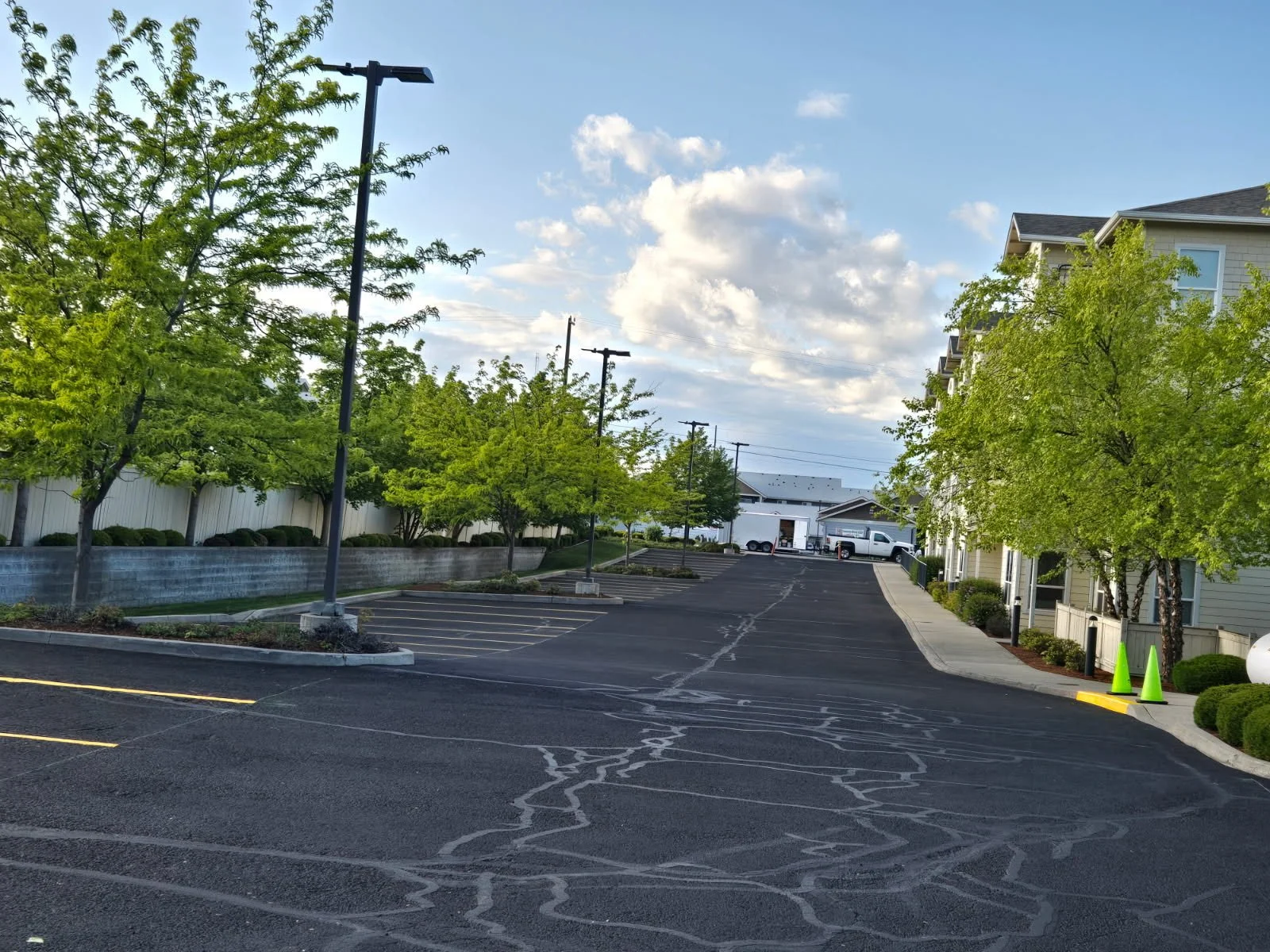 Empty parking lot with newly paved asphalt, green trees, a building on the right, and a cloudy sky.