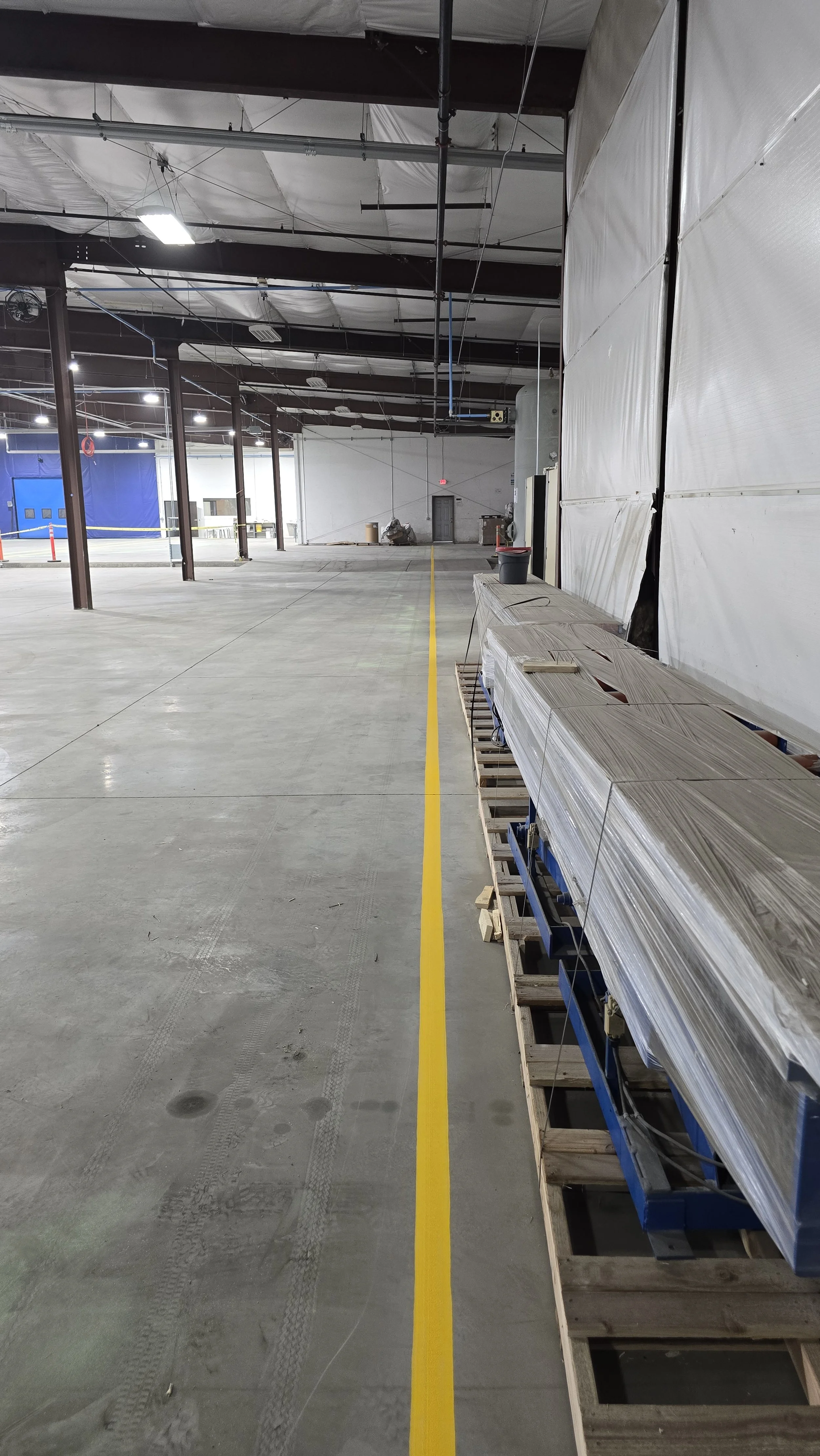 Empty warehouse or industrial space with a yellow line on the concrete floor, construction materials and temporary wall coverings along the right side, and ceiling lighting.
