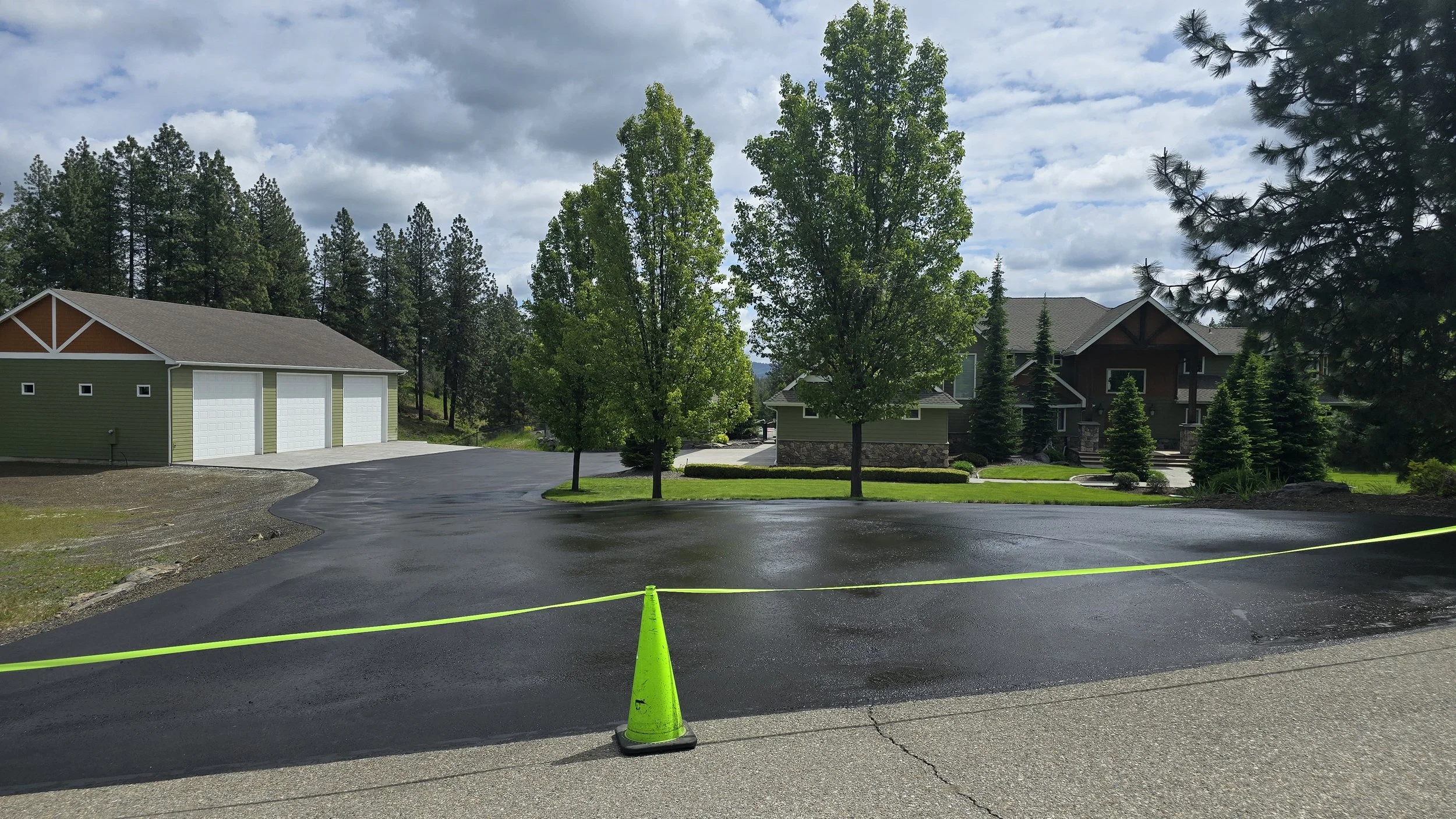 A newly paved black asphalt driveway in front of a house, cordoned off by a bright yellow traffic cone and yellow caution tape, with trees and a house in the background under a partly cloudy sky.