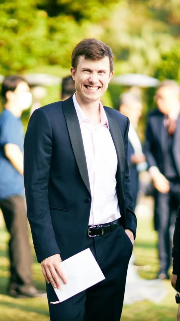 Man in a suit smiling outdoors with people in the background.