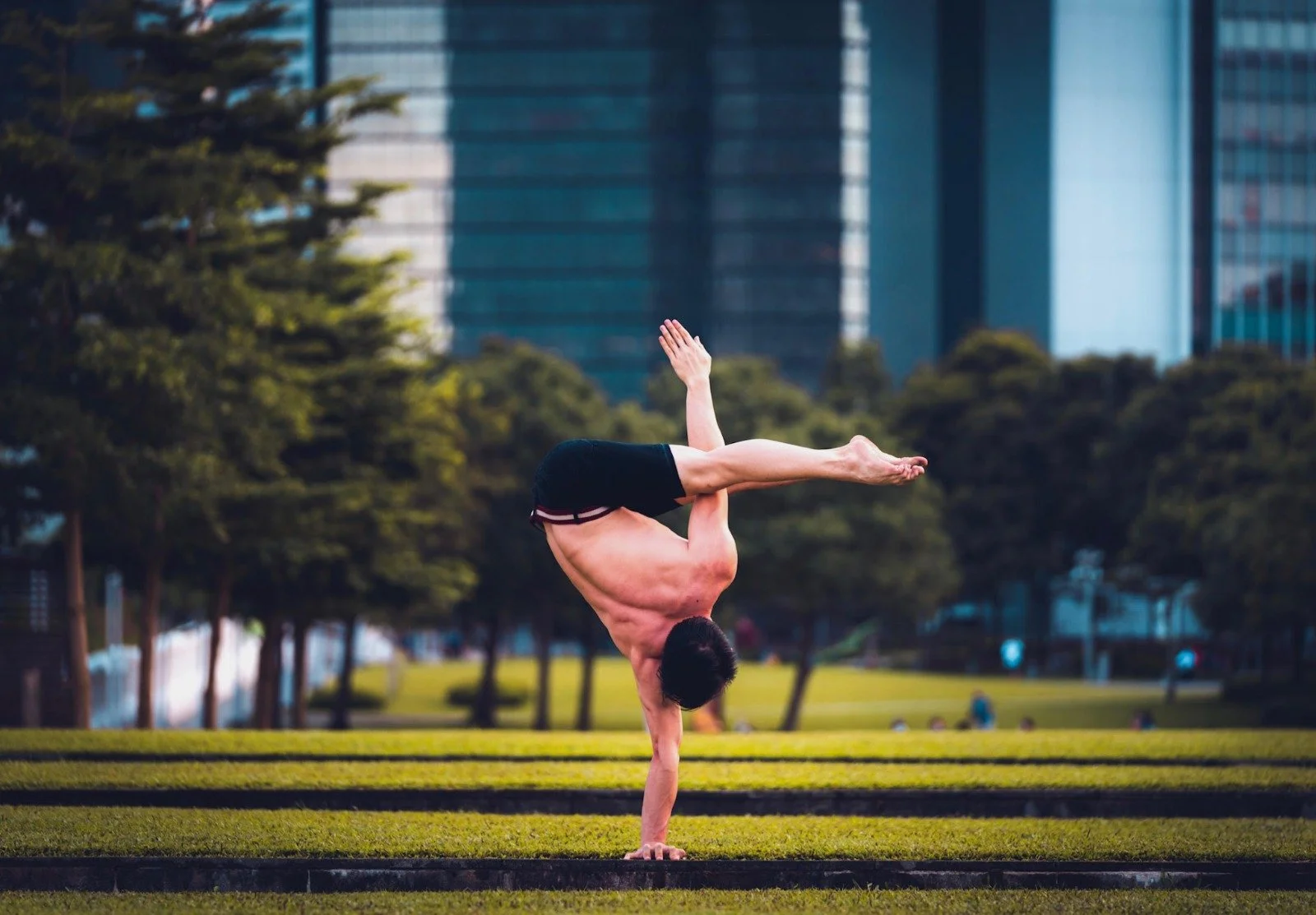 Man performing one-handed handstand outdoors with trees and buildings in the background.