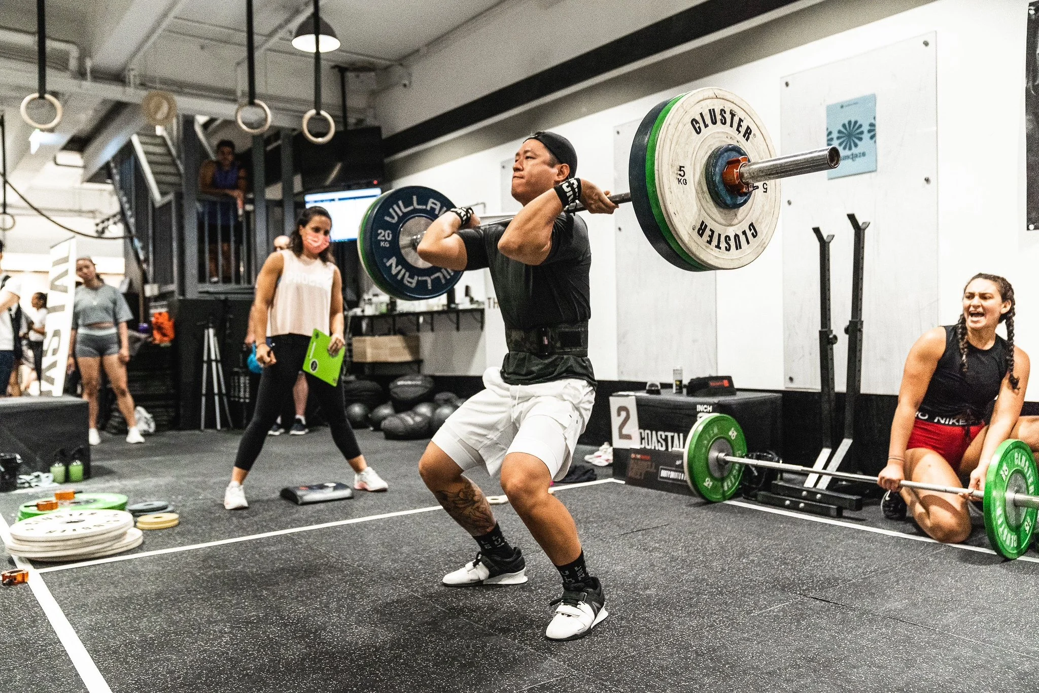 A person lifting a heavy barbell in a gym setting, surrounded by other people and gym equipment. Some are observing and taking notes, others are preparing their own weights. The atmosphere is energetic and focused.