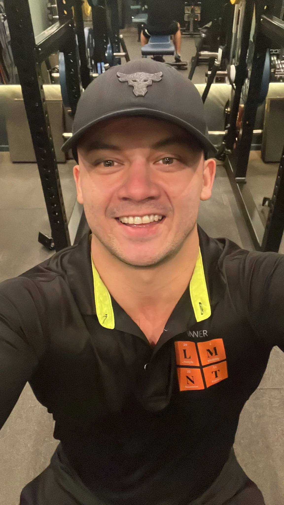 Man taking a selfie in a gym, wearing a baseball cap and black shirt with yellow accents.