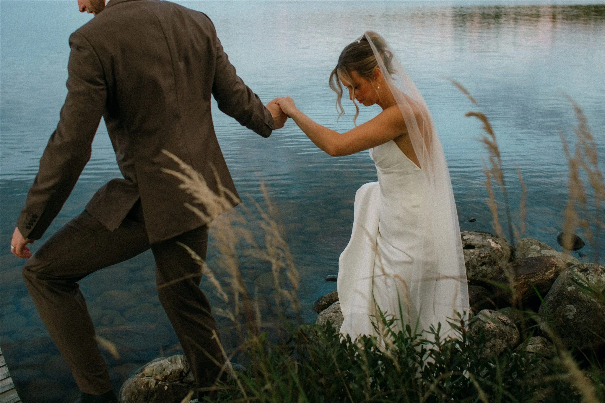 Groom helping bride off rocks at lakeside wedding at Northridge Inn