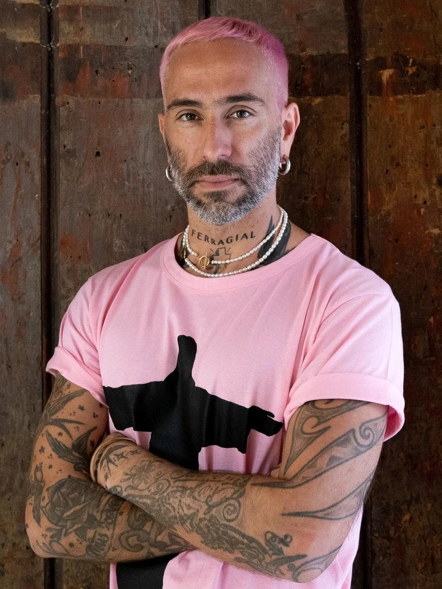 A man with pink hair, tattoos, and earrings, wearing a pink T-shirt with a black silhouette of a hand and thumb up, standing with arms crossed in front of a wooden wall.