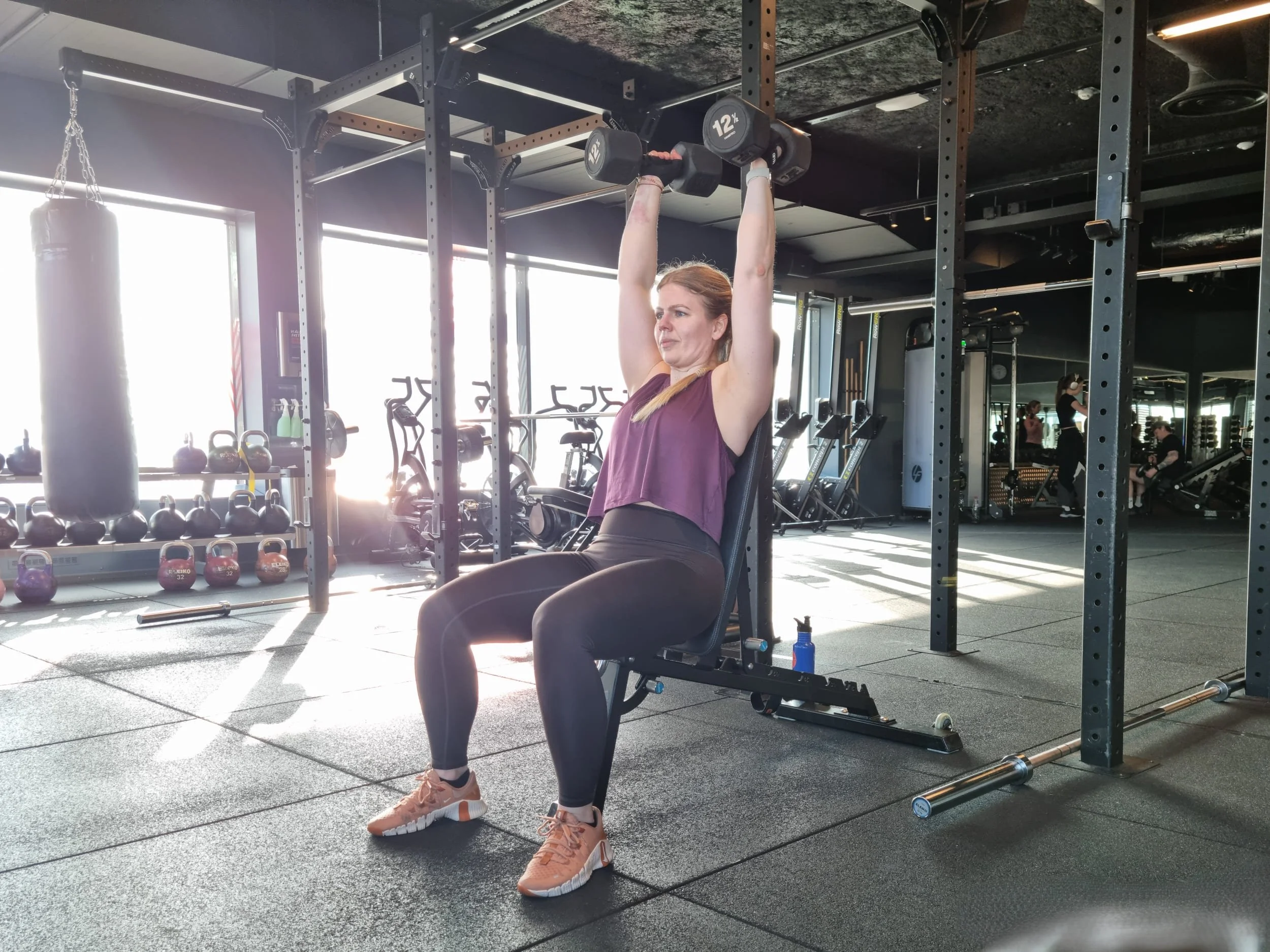 StinaP gör sittande axelpress på ett gym med solljus genom fönstren