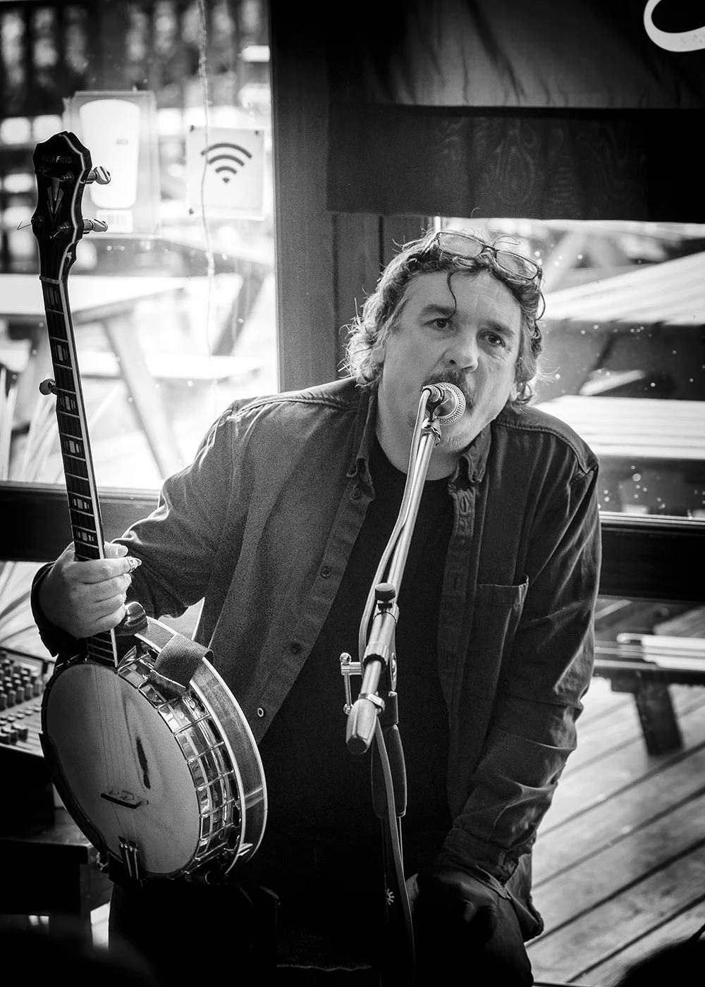 A man with curly hair and glasses on his head is singing into a microphone while holding a banjo in a room with large windows and outdoor picnic tables visible outside.