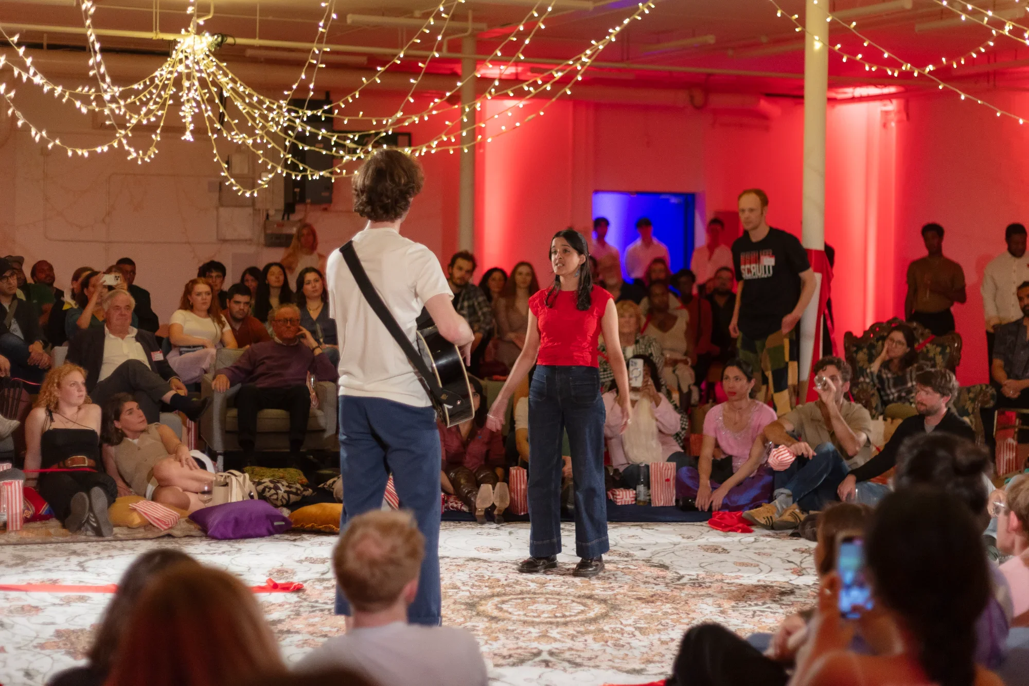 The SCRUM Company in performance in the Warehouse space. A packed house watch a man with a guitar and a woman singing — another performer walks towards them. You could hear a pin drop.