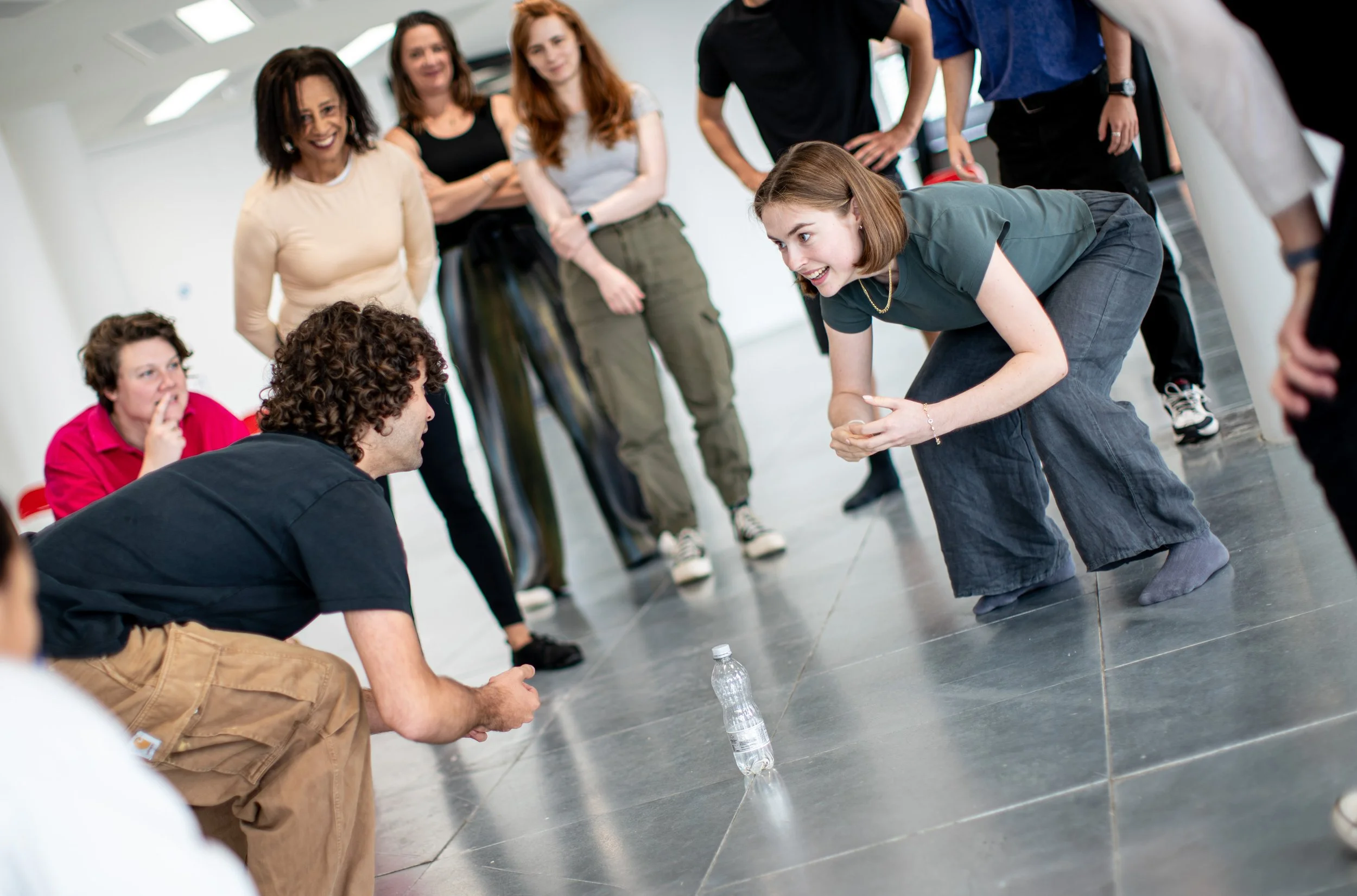 Two actors crouching, poised over a bottle of water. Everyone in the room seems very excited about who's going to grab it first. From The Shake Up Intensive, July 2025.