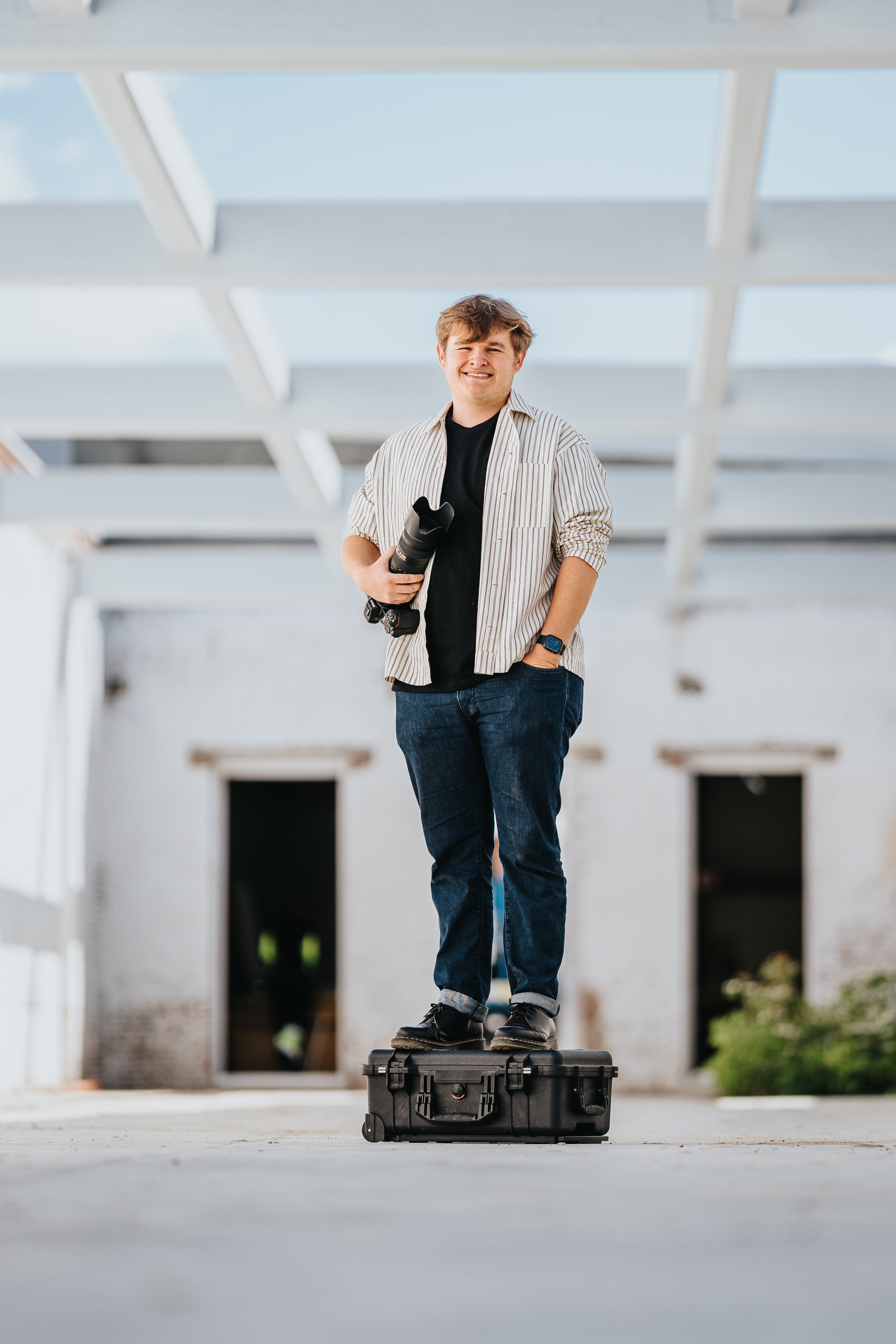 A young man standing outdoors on a black protective case, holding a camera lens with a smile.