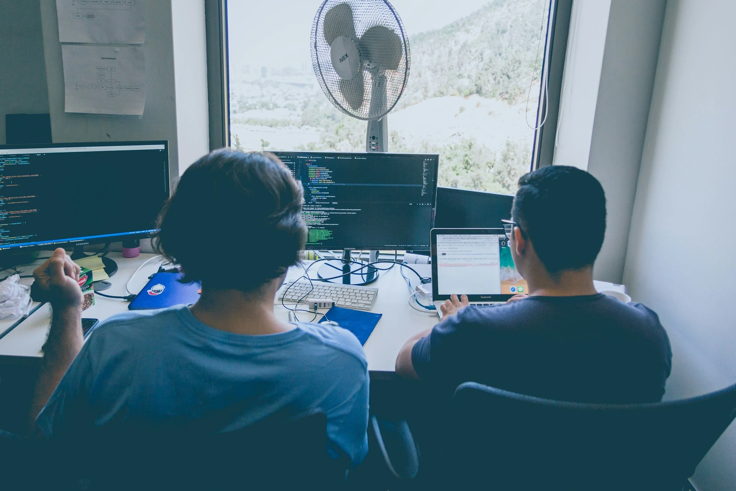 Two people working at a desk with multiple monitors, a tablet, and a fan near a window with a view of hills and trees.