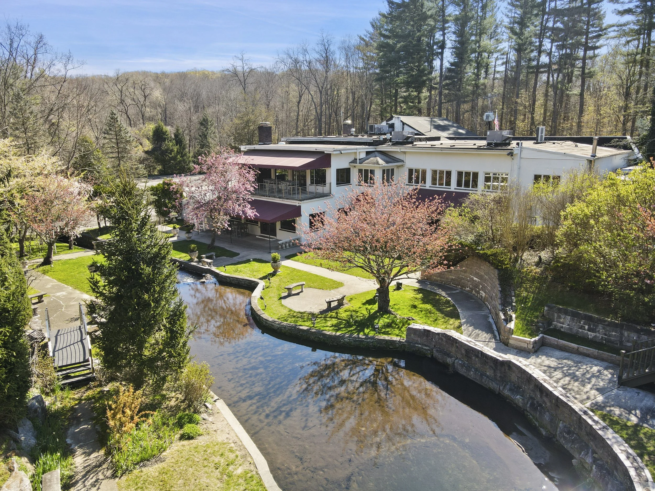 Aerial view of a white building with a maroon awning, surrounded by blooming cherry blossom trees, a pond, benches, and a pathway in a landscaped garden area with forest in the background.
