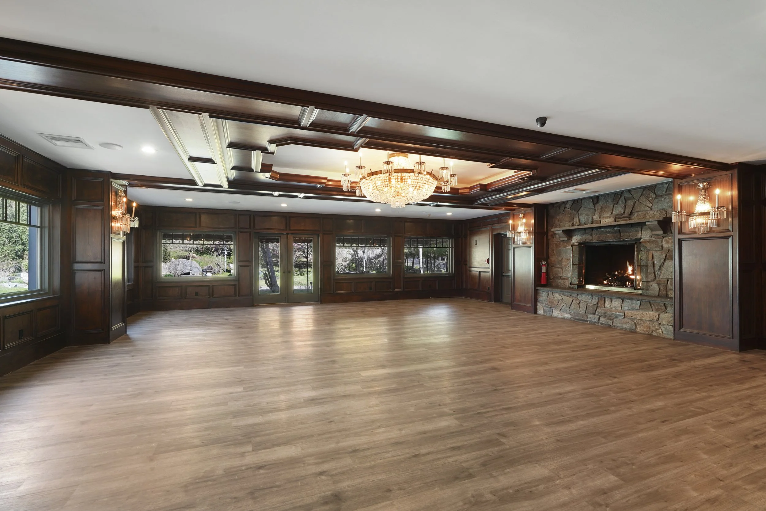 Spacious room with hardwood floors, wood-paneled walls, a stone fireplace, and a large chandelier hanging from the ceiling.