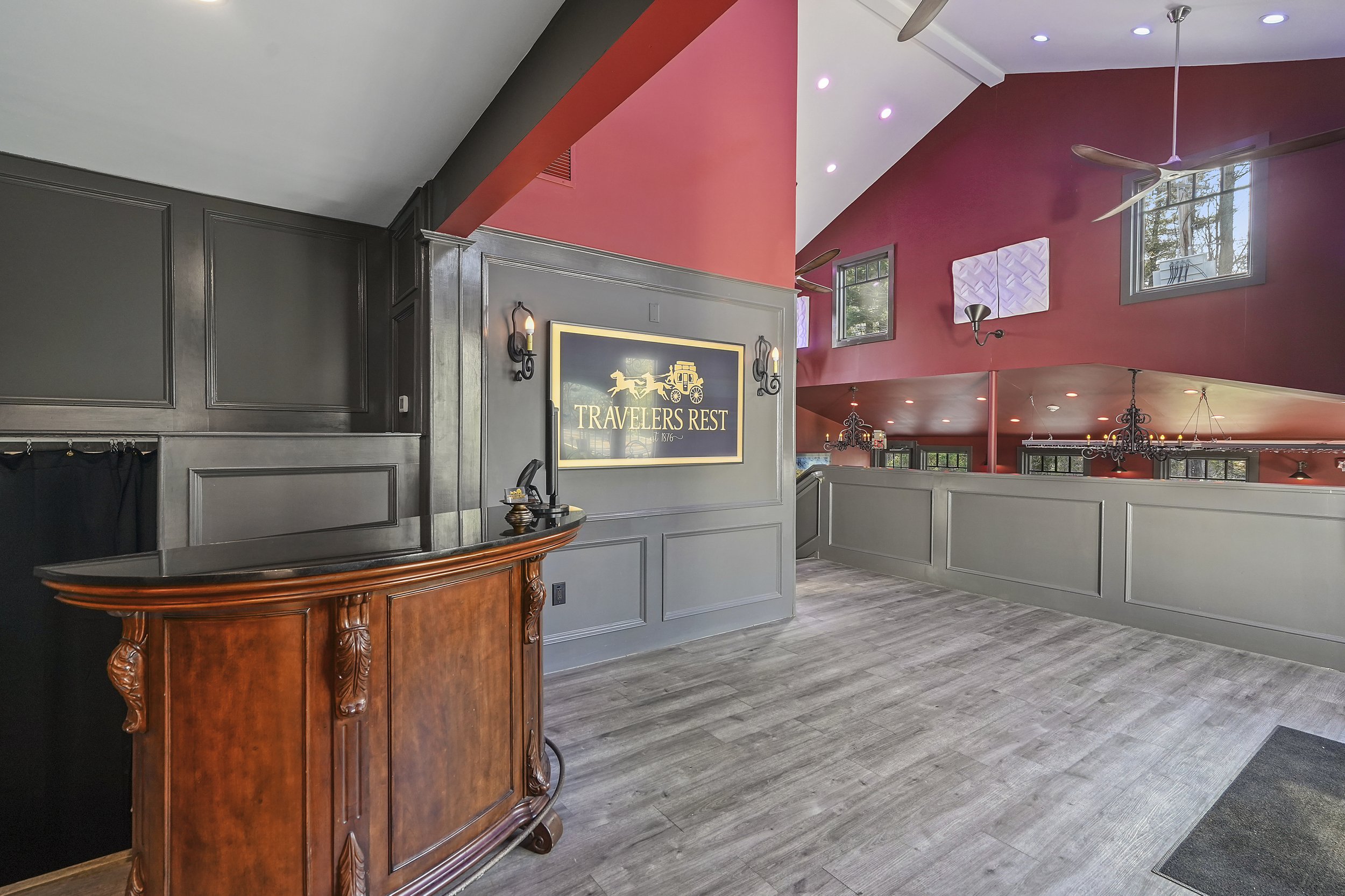 Interior of a restaurant named 'Travelers Rest' with a vintage wooden reception desk, gray wooden paneling, a large wall sign, and a high ceiling with fans, chandeliers, and multiple windows with natural light.
