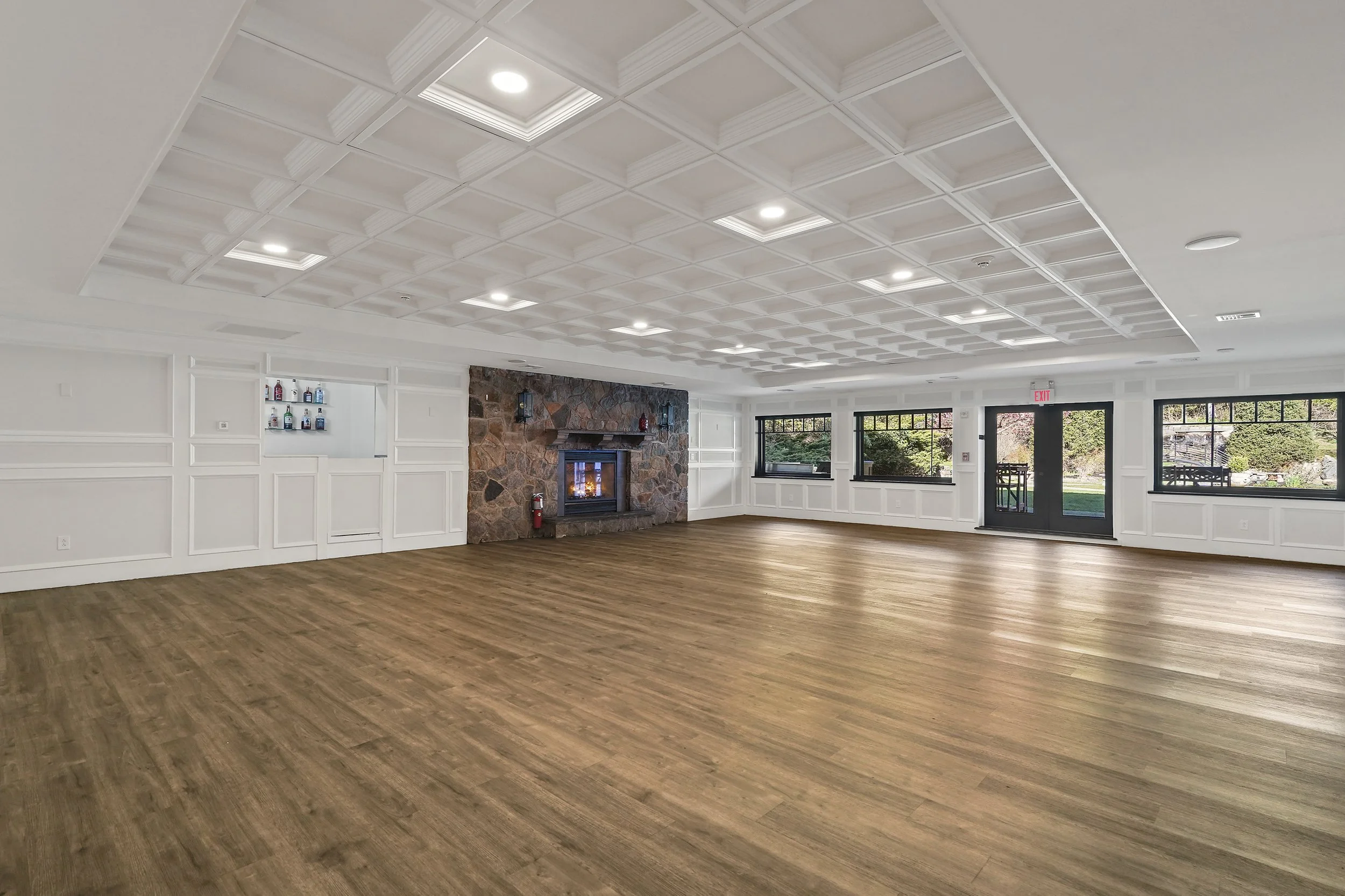 Empty room with wood flooring, white paneled walls, large windows, a stone fireplace, and a ceiling with recessed lighting.