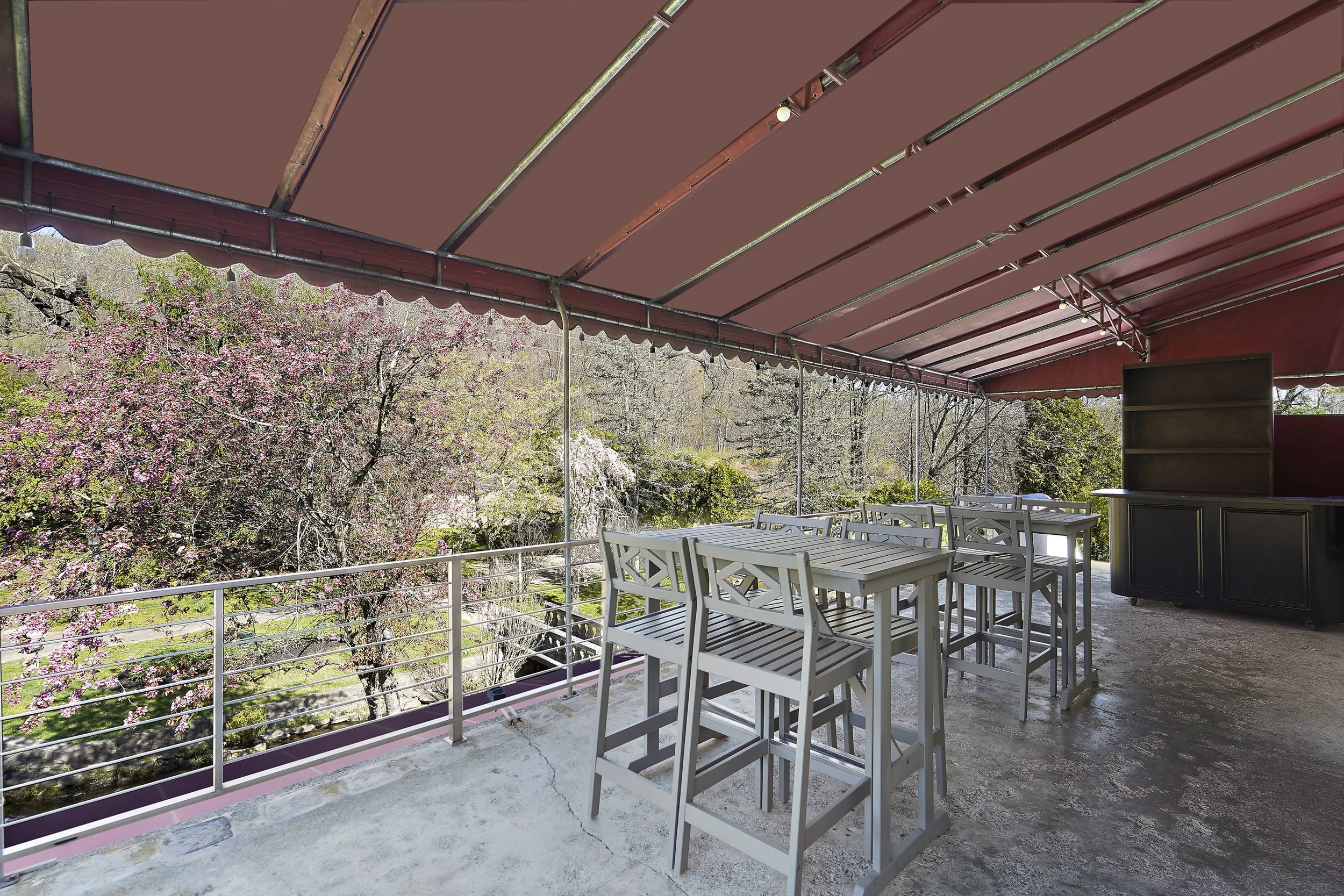 An outdoor patio with white wooden tables and chairs under a maroon canopy, overlooking a garden with trees and blooming pink flowers.