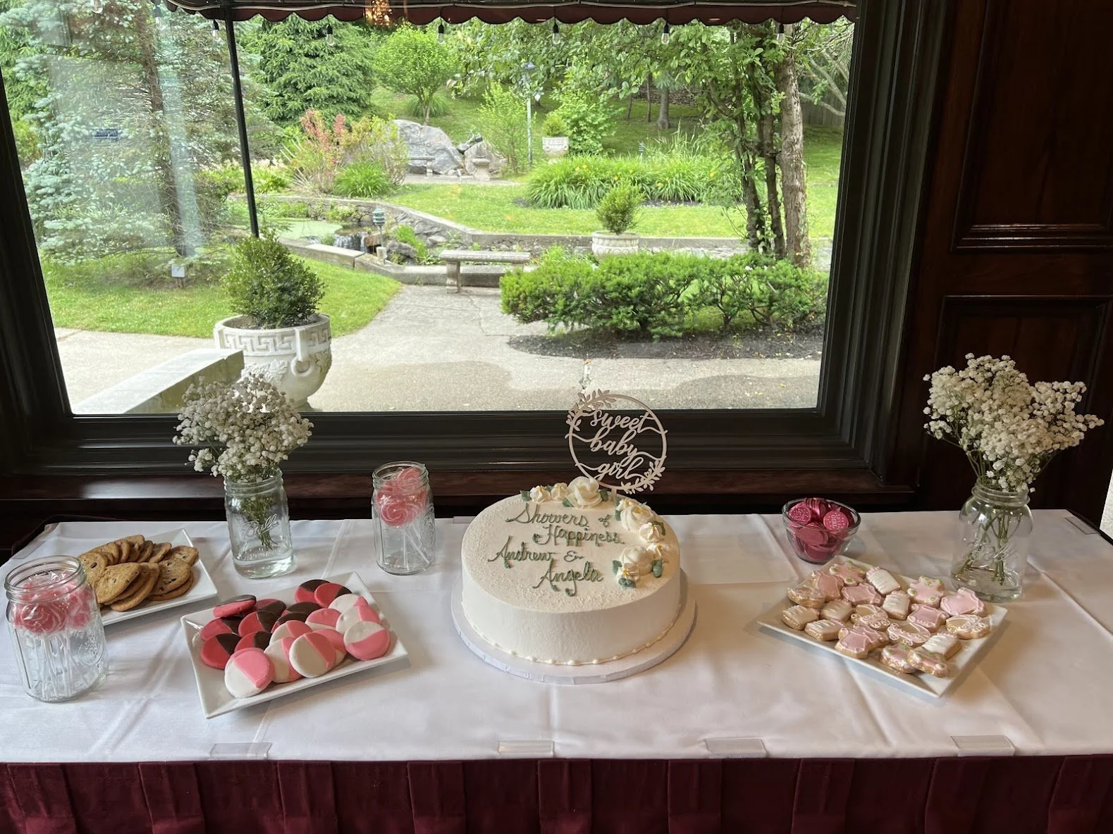 A table with desserts and a birthday cake in front of a window overlooking a garden. The table has a white tablecloth, two glass vases with white baby's breath flowers, mason jars with pink and white feather decorations, plates of cookies, pink and w