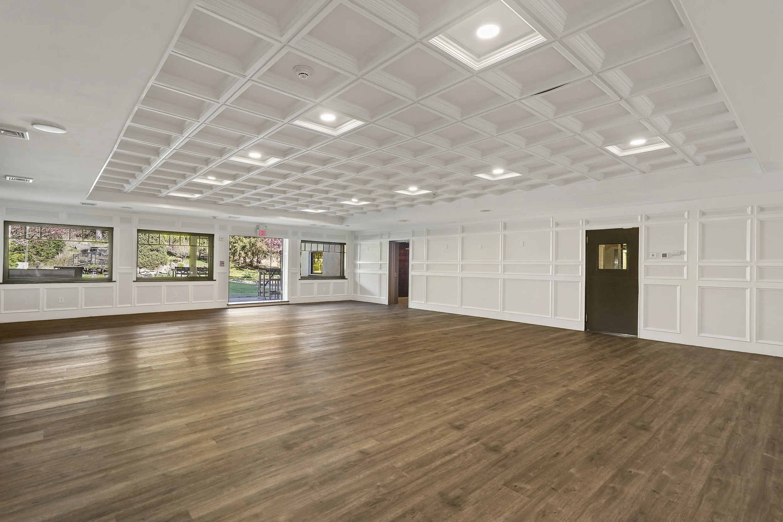 Empty spacious room with white paneled walls, large windows, a glass door, and a wood floor.