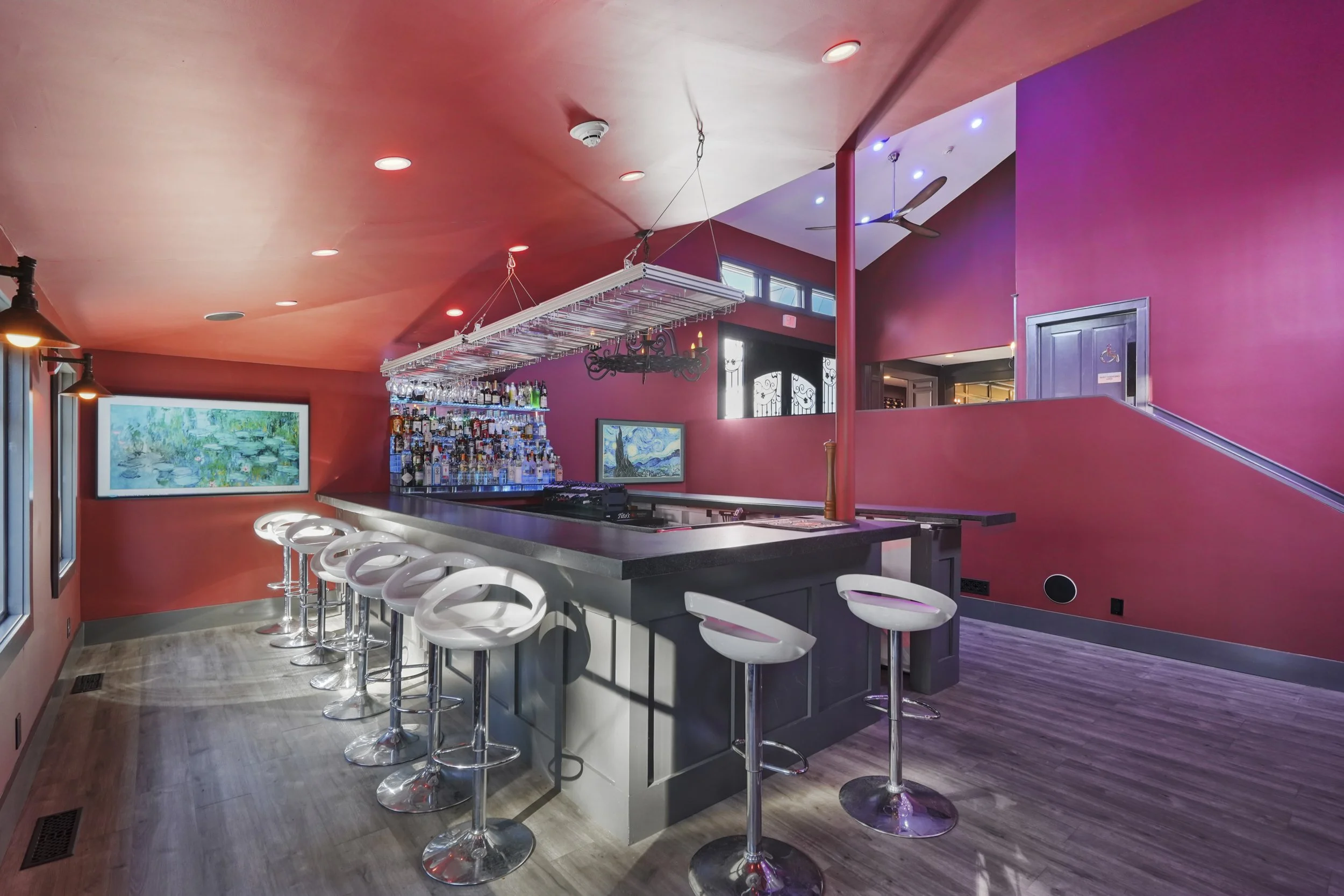 A modern bar with a curved counter, white bar stools, a ceiling-mounted rack with hanging glasses, a liquor shelf behind the bar, and vibrant pink walls with artwork. There are large windows on the left and blue accent lighting.