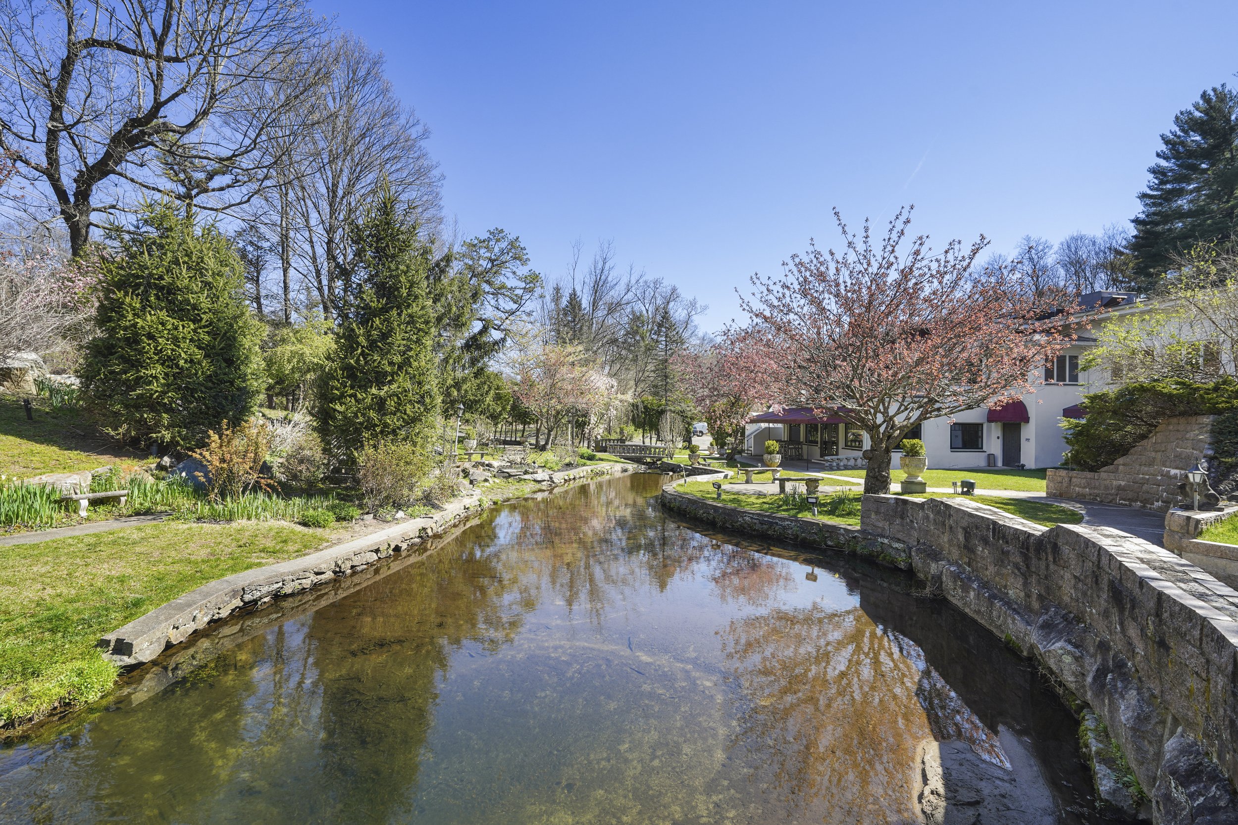 A peaceful outdoor scene with a small stream bordered by a stone wall, surrounded by blossoming trees and lush green grass, with a house featuring red awnings in the background under a clear blue sky.