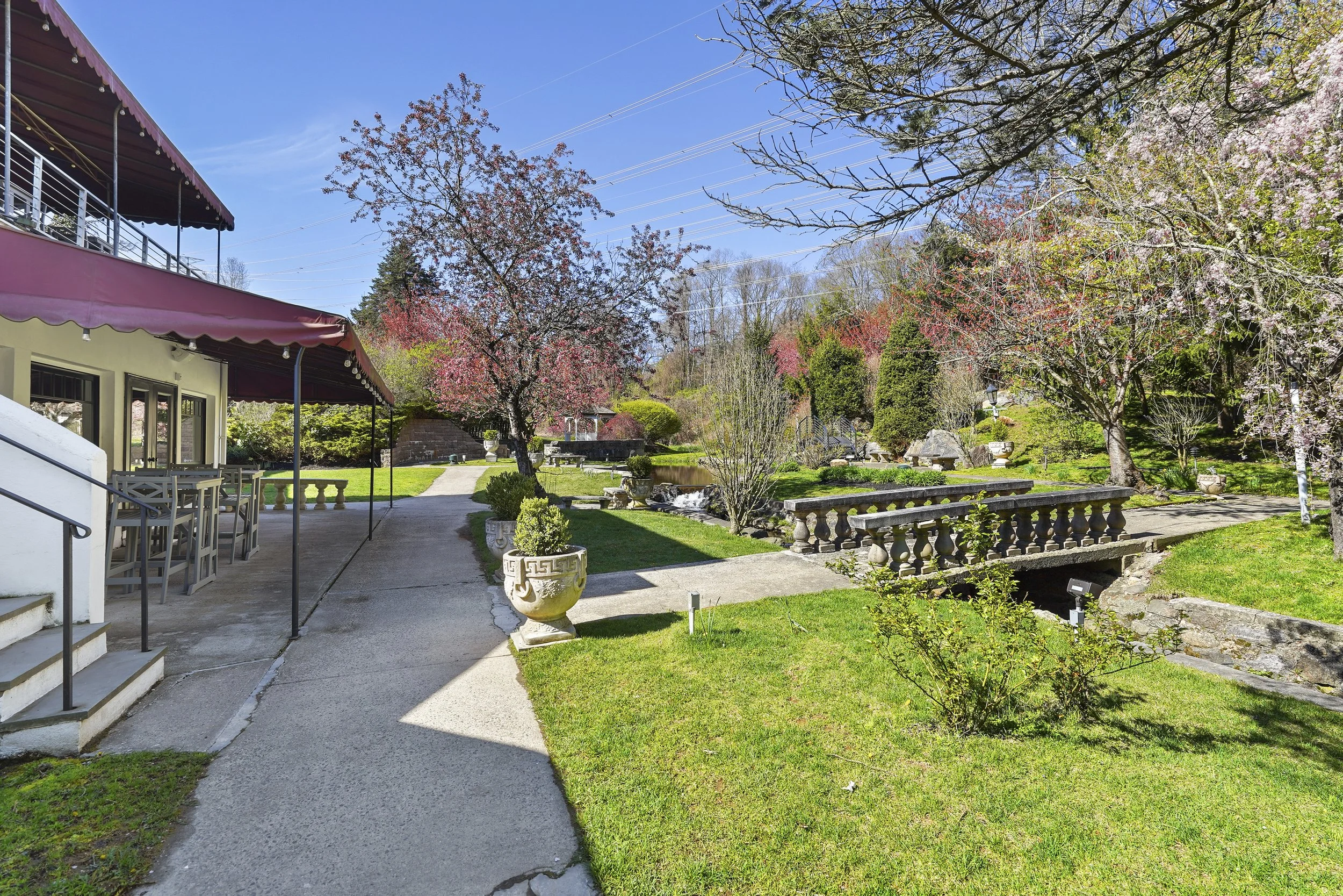A lush garden with blooming pink and white cherry blossom trees, a paved walkway, potted plants, decorative stone railings, and a house with a red awning under a blue sky.