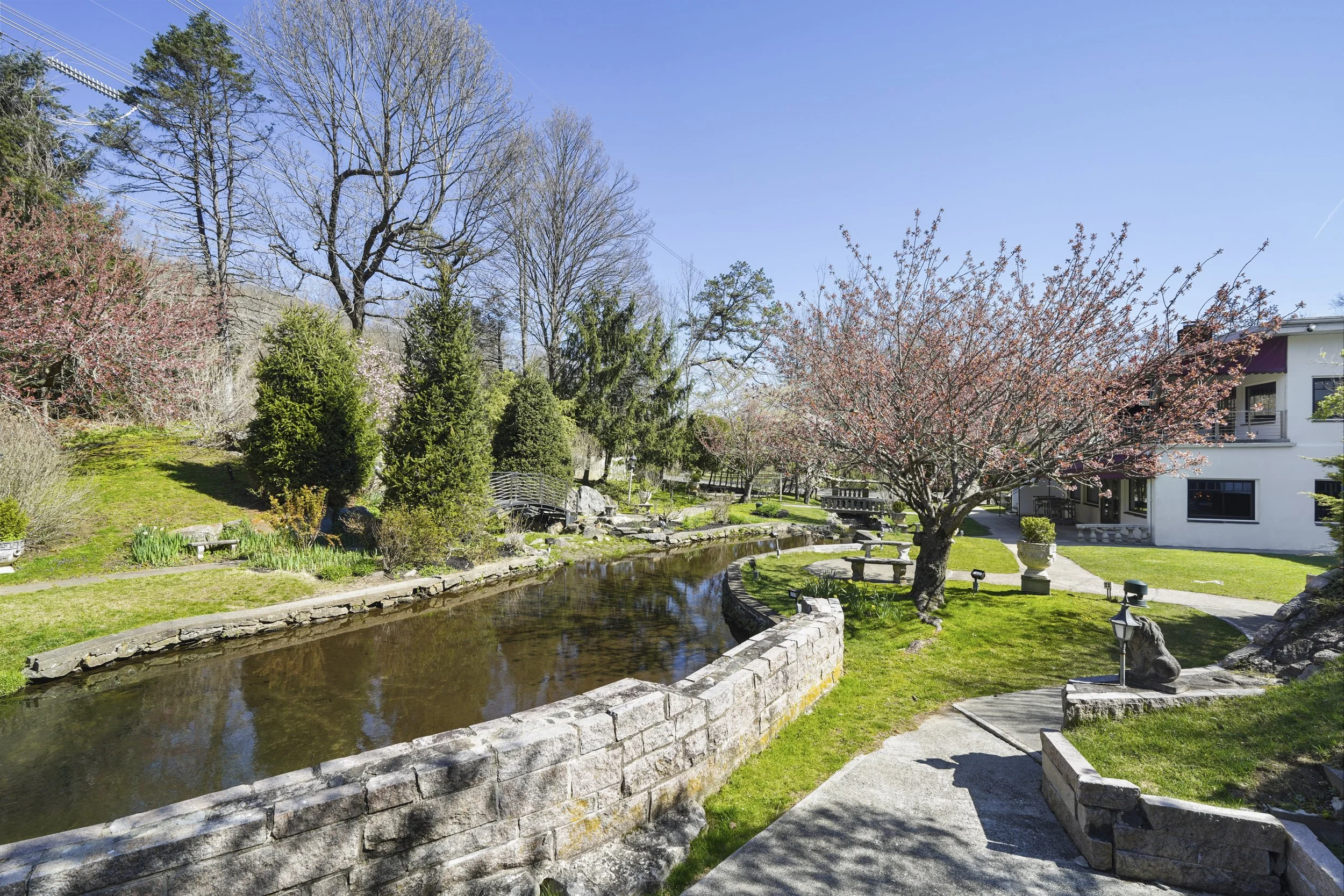 A landscaped garden with a small pond, a tree with pink blossoms, various bushes and trees, a stone wall around the pond, benches, and a white building in the background.