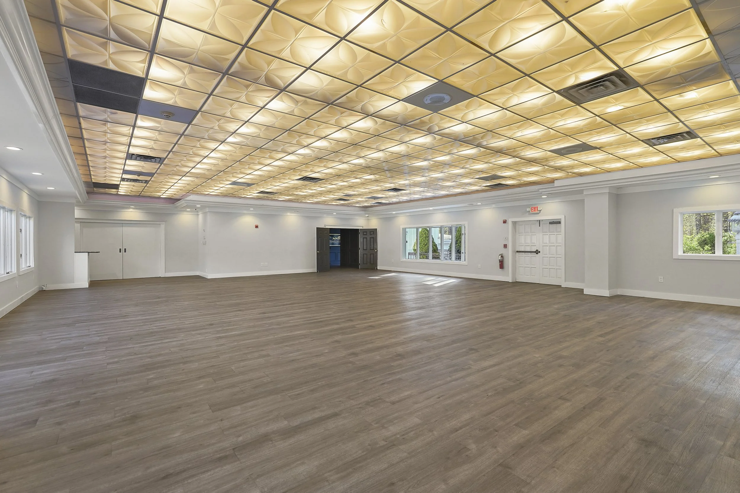 Large empty event hall with wooden flooring, white walls, and a decorative ceiling with a grid pattern of illuminated panels. Multiple windows and a double door are visible.