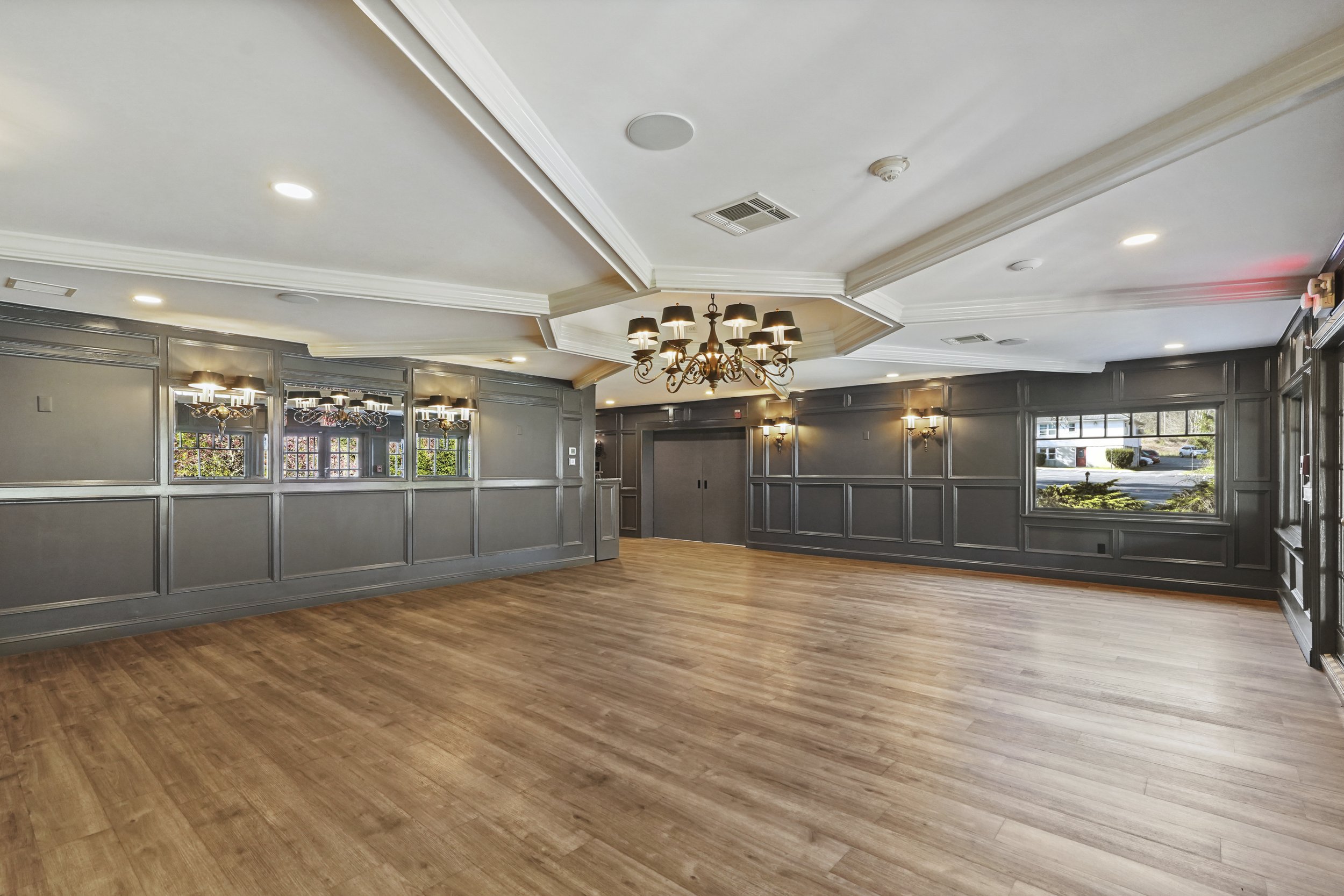 Large, empty room with hardwood floor and gray paneled walls, illuminated by chandeliers and wall sconces, with windows showing outdoor scenery.