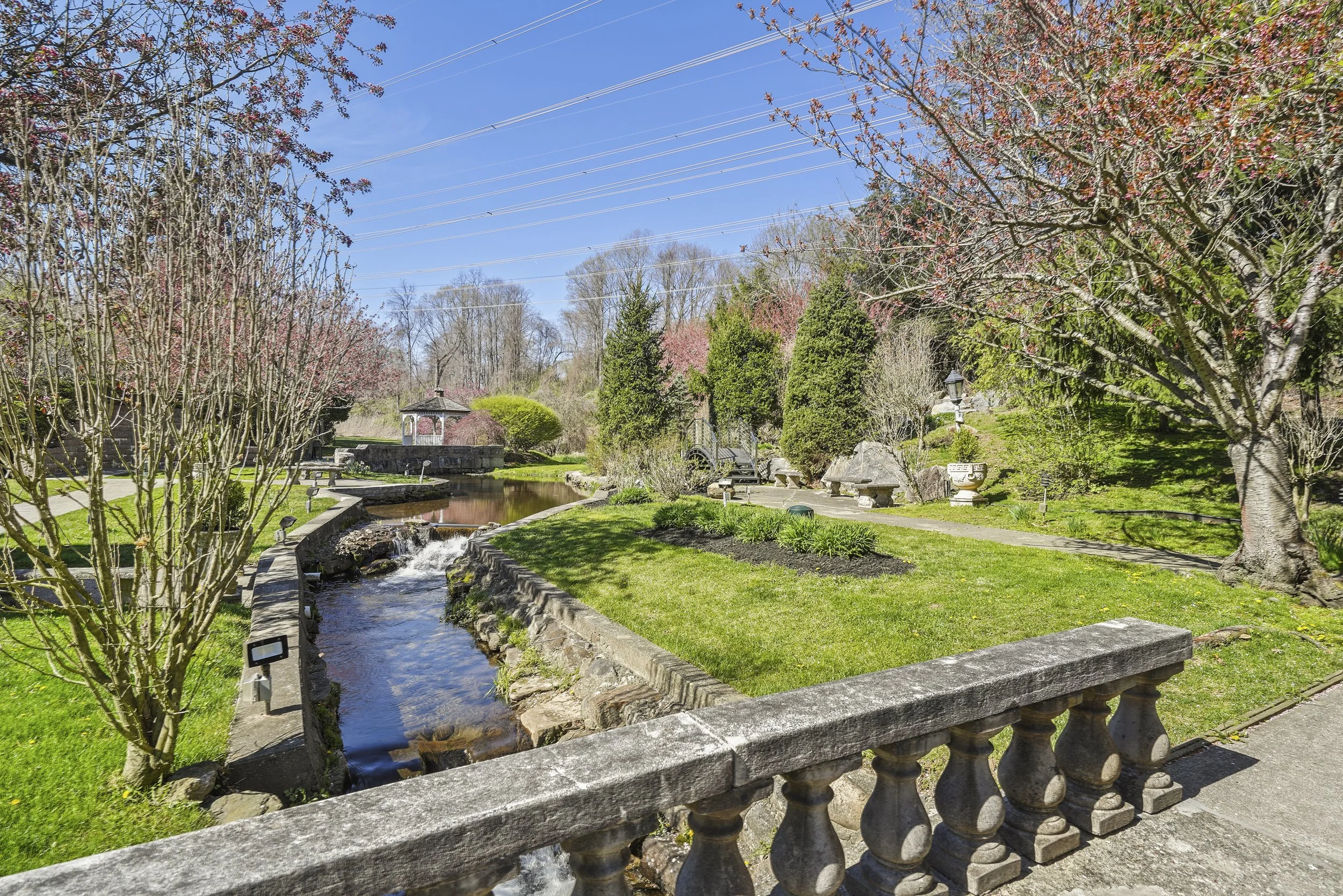 A scenic park featuring a small flowing stream bordered by stone and concrete, surrounded by green grass, trees, and shrubs with pink blossoms, with a gazebo in the background and a blue sky overhead.