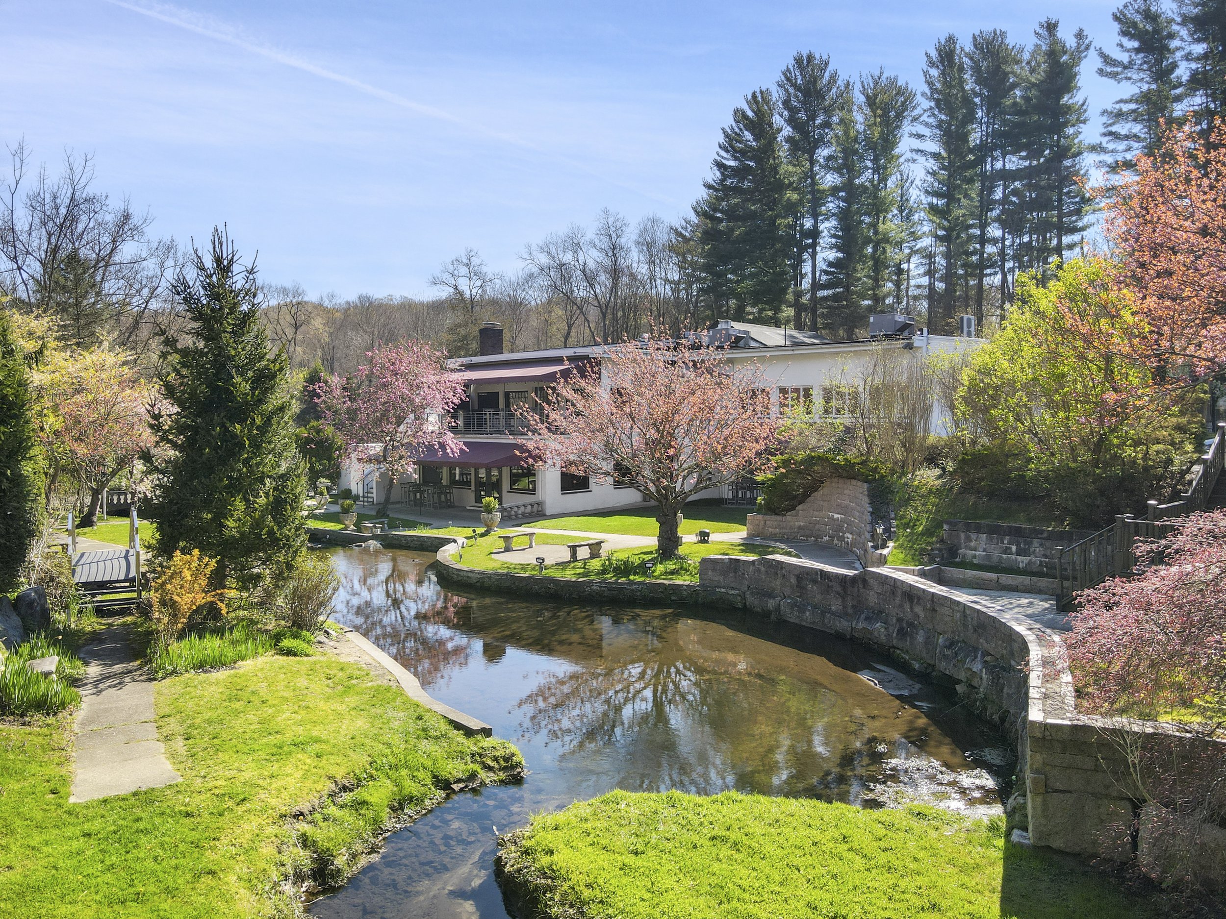 A large house with a garden and a pond, surrounded by trees with pink blossoms on a clear sunny day.