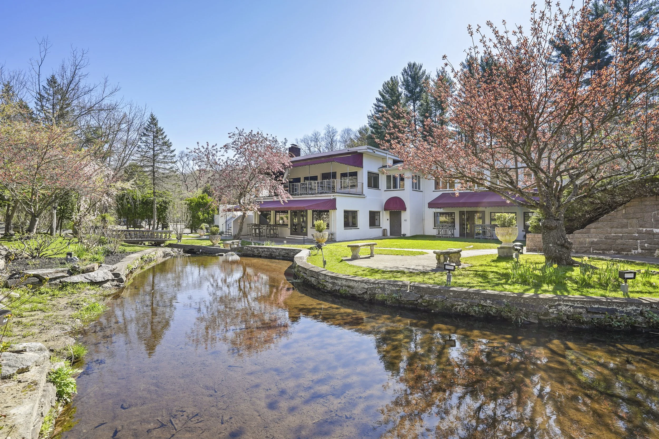 A house with pink awnings and balconies, surrounded by blossoming trees, is situated near a small pond reflecting the clear blue sky.