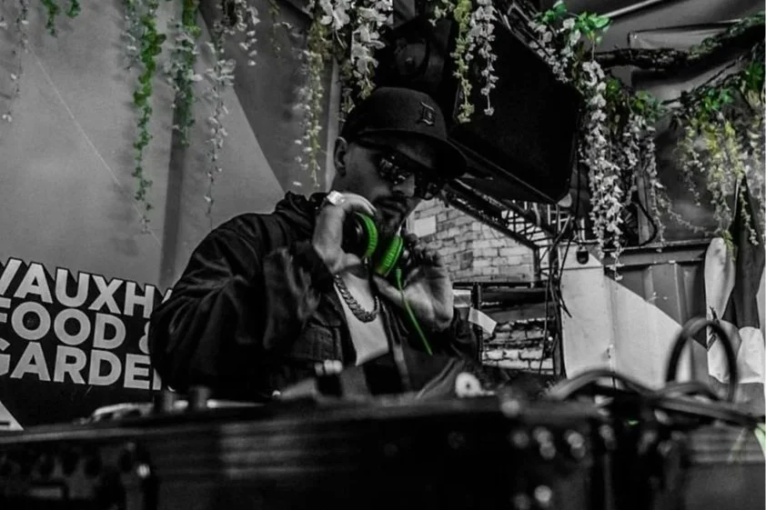 A DJ wearing sunglasses, a baseball cap, and a chain, holding headphones in one hand in a decorated venue with hanging plants, bricks, and a sign that reads 'Vauxhall Food Garden'.