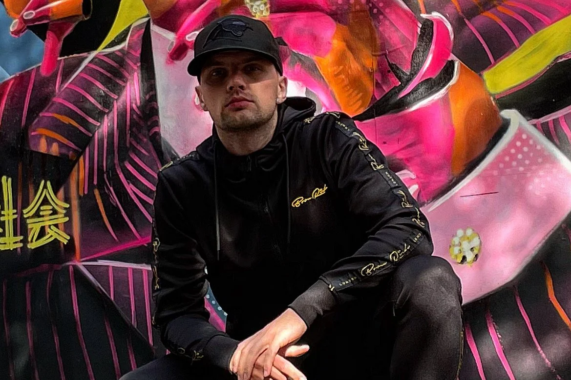 A man wearing a black cap and black hoodie sitting in front of a colorful graffiti mural with pink, orange, green, and yellow elements.