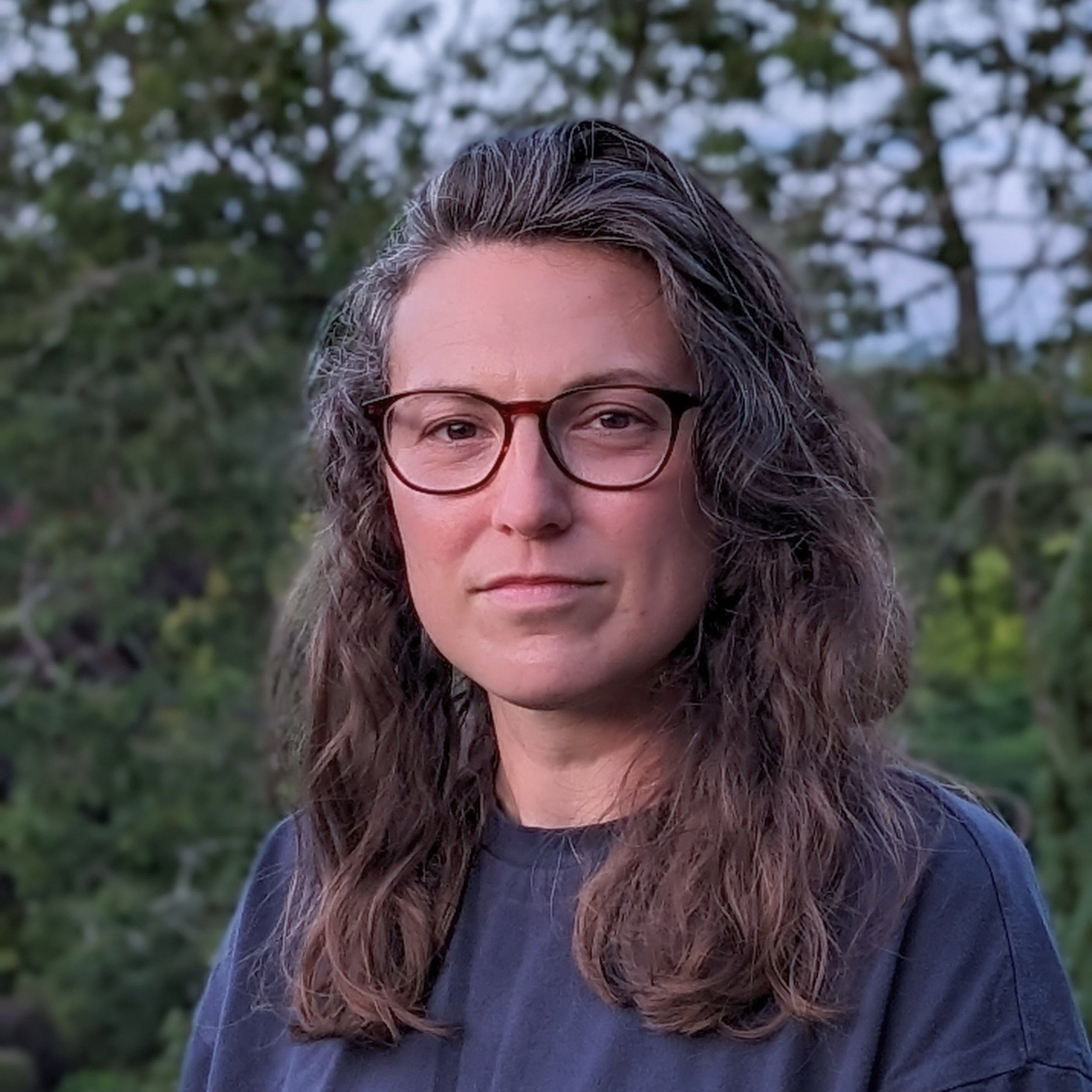 A woman with wavy brown hair, wearing glasses and a dark blue shirt, standing outdoors with trees in the background.
