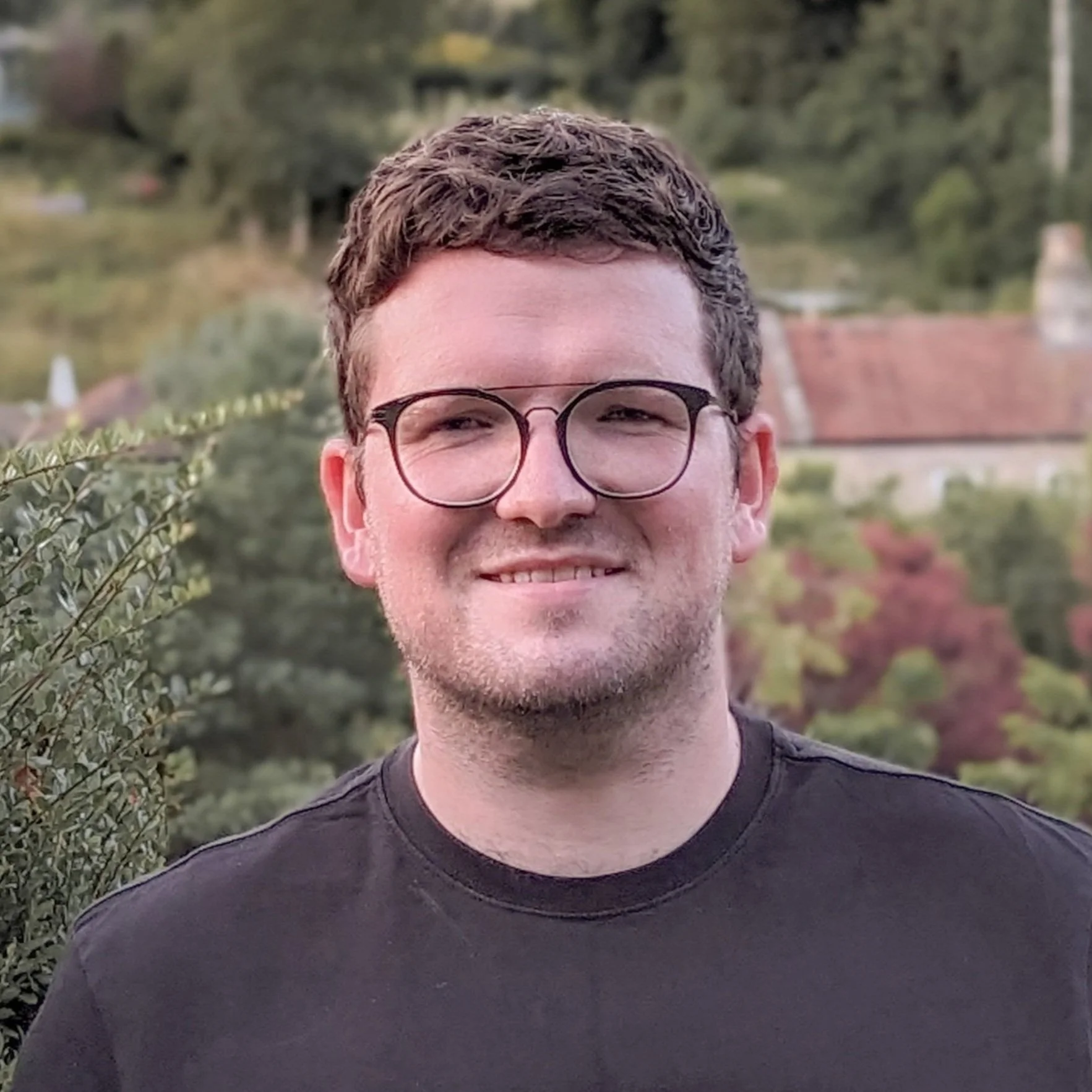 Young man with glasses and short brown hair smiling outdoors, with trees and houses in the background.