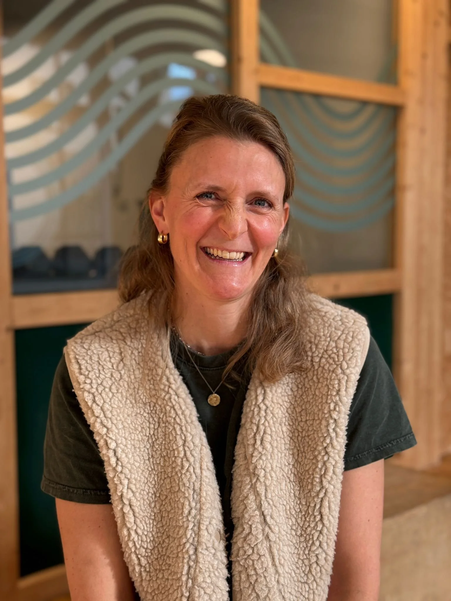 Eine lachende Frau mit brauen, gewellten Haaren, trägt goldene Ohrringe und eine Halskette. Sie trägt ein schwarzes Shirt und eine beige, wollige Weste. Im Hintergrund sind Holzmöbel und ein dekoratives Fenster zu sehen.